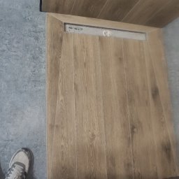 Polwykon - Ukończona podłoga w łazience z drewnopodobnych płytek, z widocznym odpływem liniowym. Szare płytki na ścianie i podłodze. Fragment buta w lewym dolnym rogu.