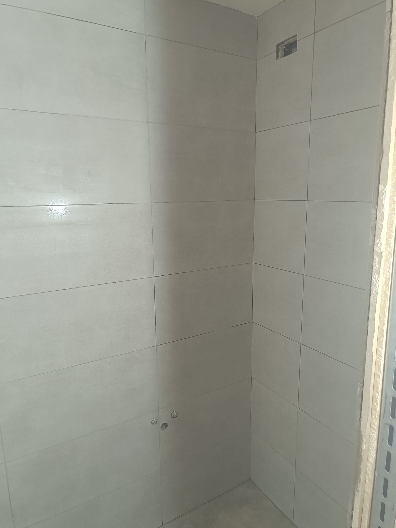 Wykończona ściana w łazience z jasnoszarymi, prostokątnymi płytkami. Widoczne otwory na armaturę i otwór na wentylację. Minimalistyczny design.