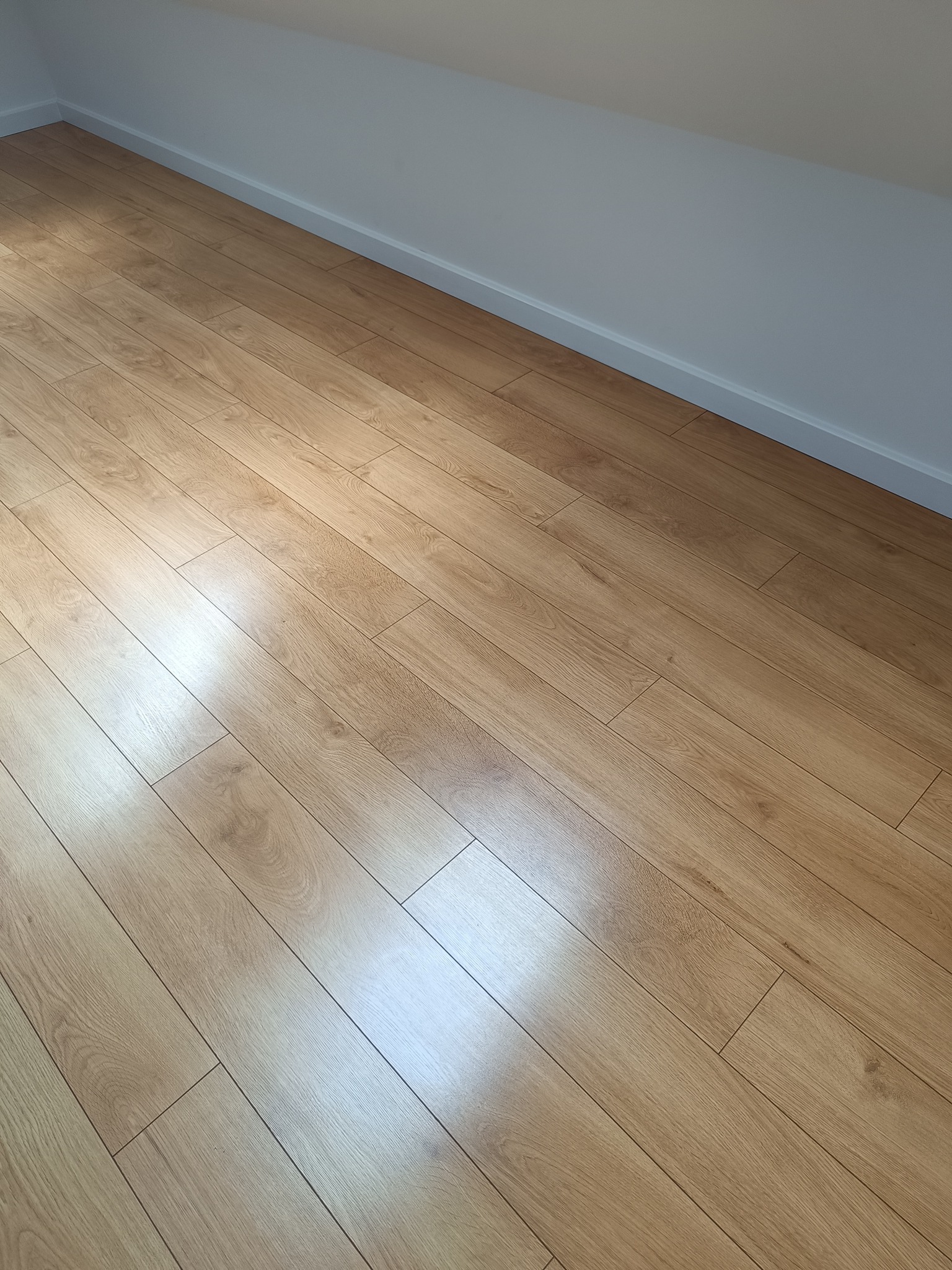 Ukończona podłoga z paneli w odcieniach jasnego drewna, ułożona w skos, z widoczną białą listwą przypodłogową i fragmentem skośnej ściany poddasza.