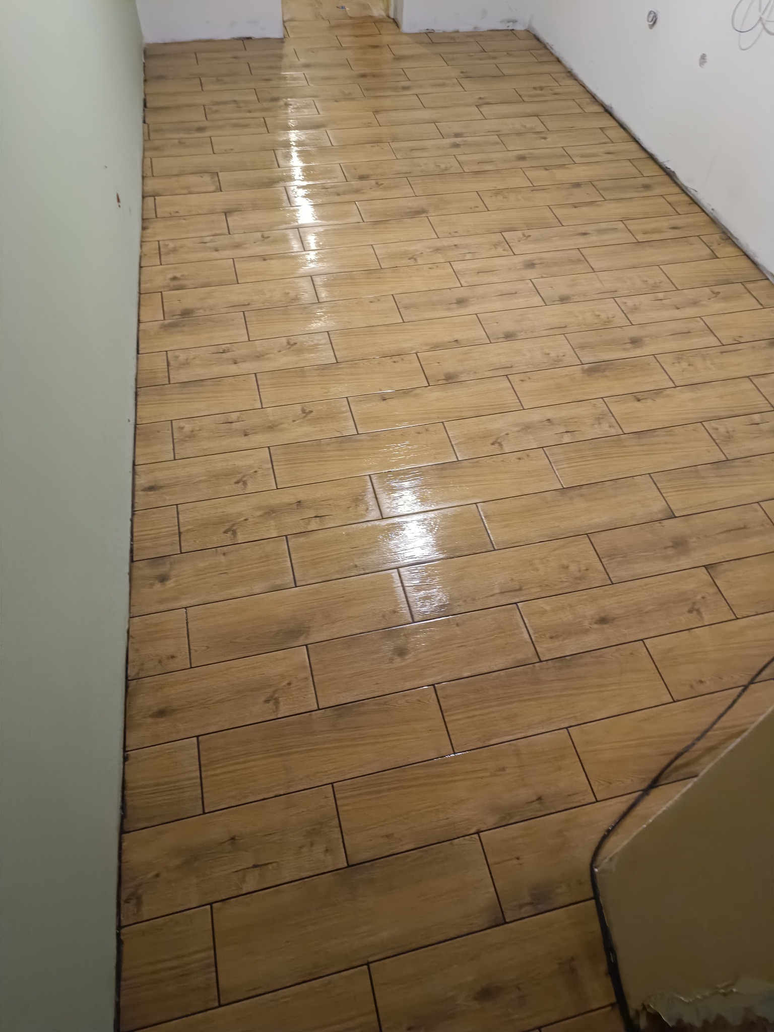 Ułożona podłoga z płytek imitujących drewno w jasnym odcieniu brązu, widok z góry, świeżo po umyciu, z połyskiem wody na powierzchni. Widoczne fragmenty ścian.