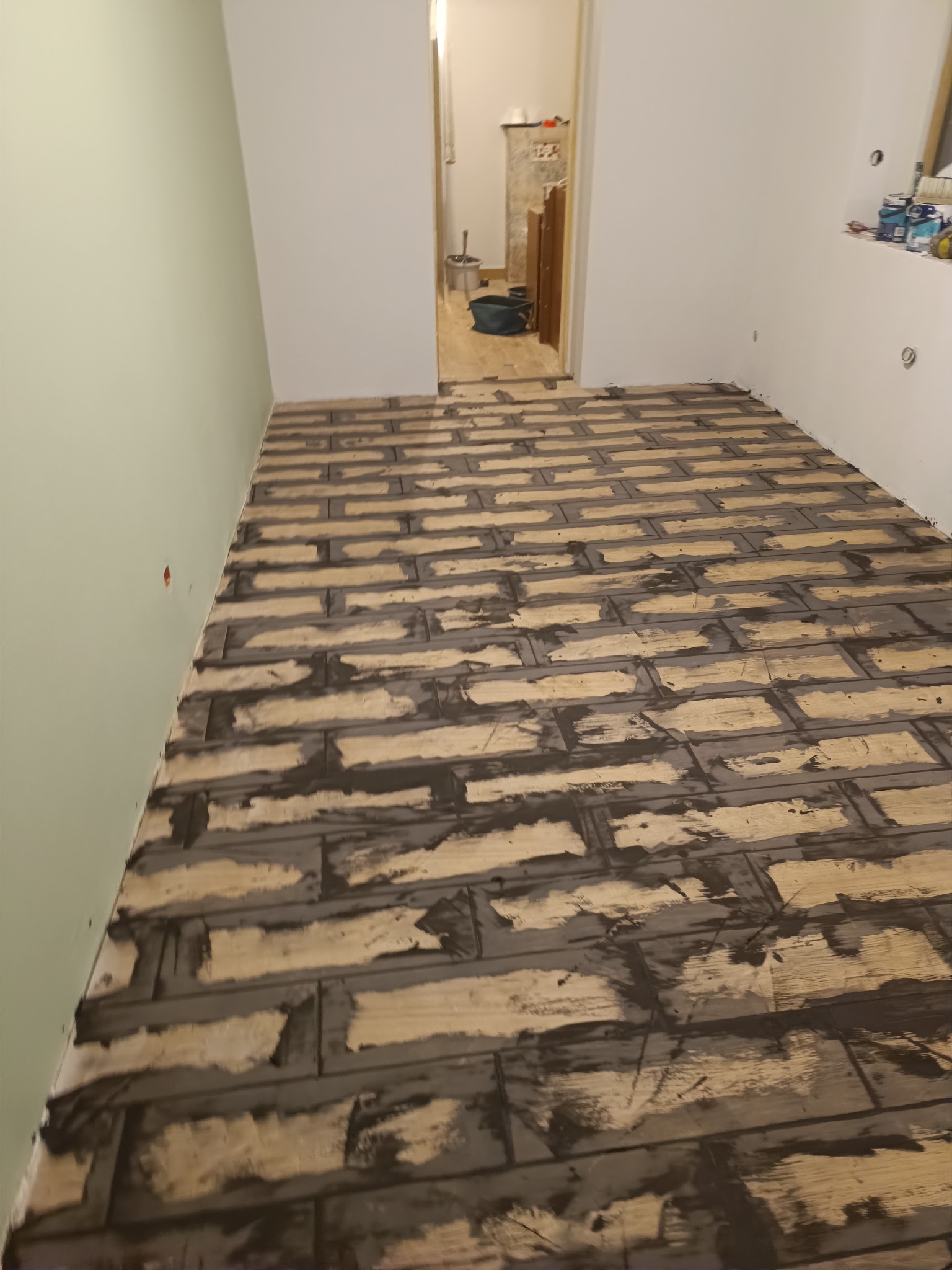 Układanie drewnianej podłogi w jodełkę, z widocznymi śladami kleju, w jasnym pomieszczeniu z zieloną ścianą. Widok z perspektywy podłogi.