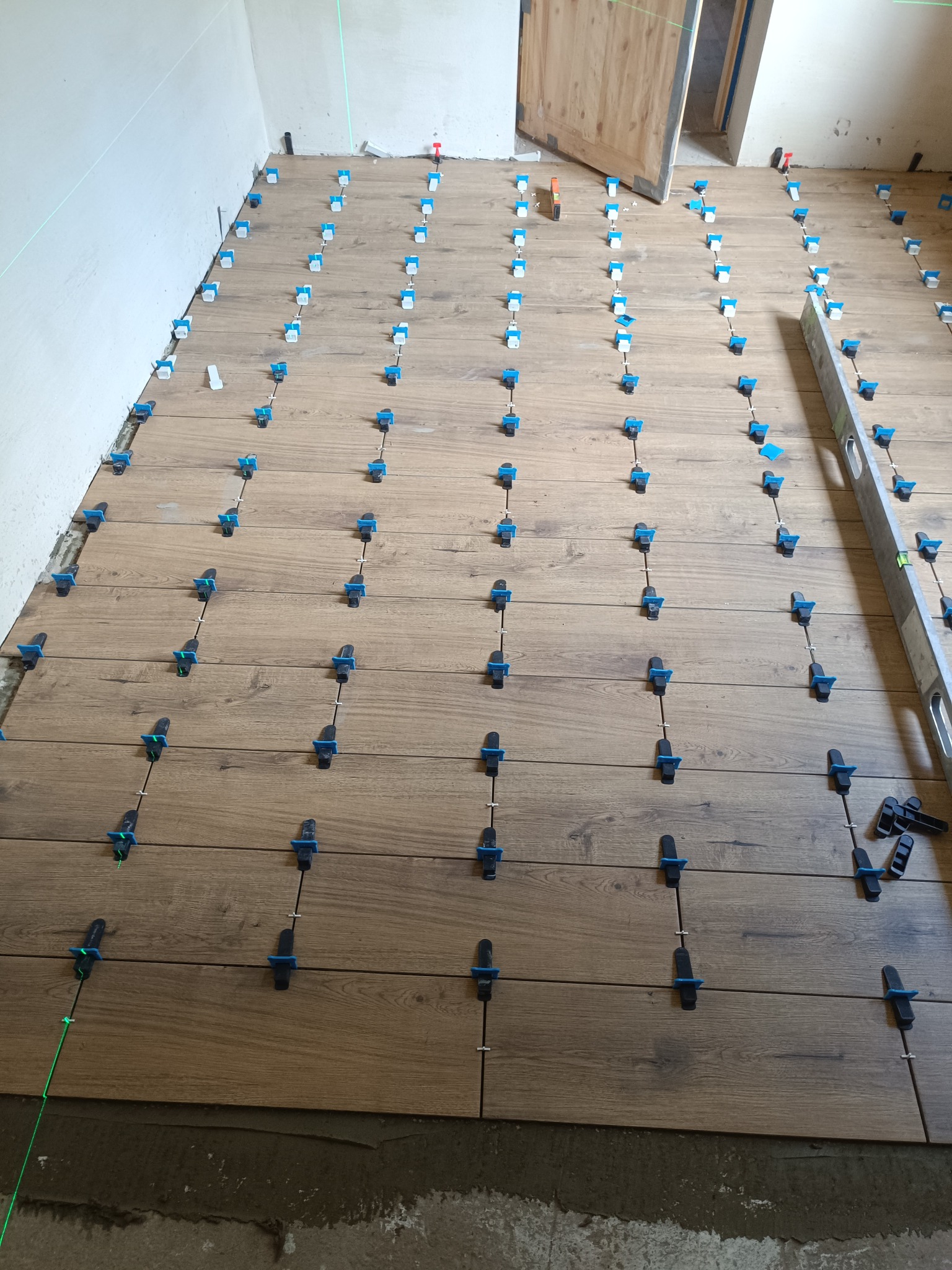 Układanie podłogi z płytek imitujących drewno z użyciem systemu poziomowania, widoczny laser krzyżowy i poziomica. Prace wykończeniowe w trakcie realizacji.