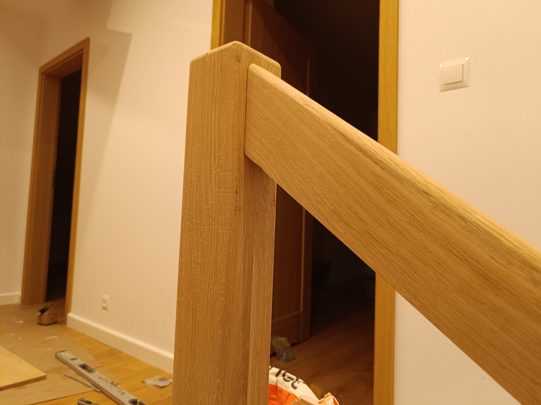 Fragment drewnianej balustrady schodowej z widocznym słupkiem i poręczą, w tle drzwi i fragment podłogi w trakcie montażu. Ciepłe oświetlenie.