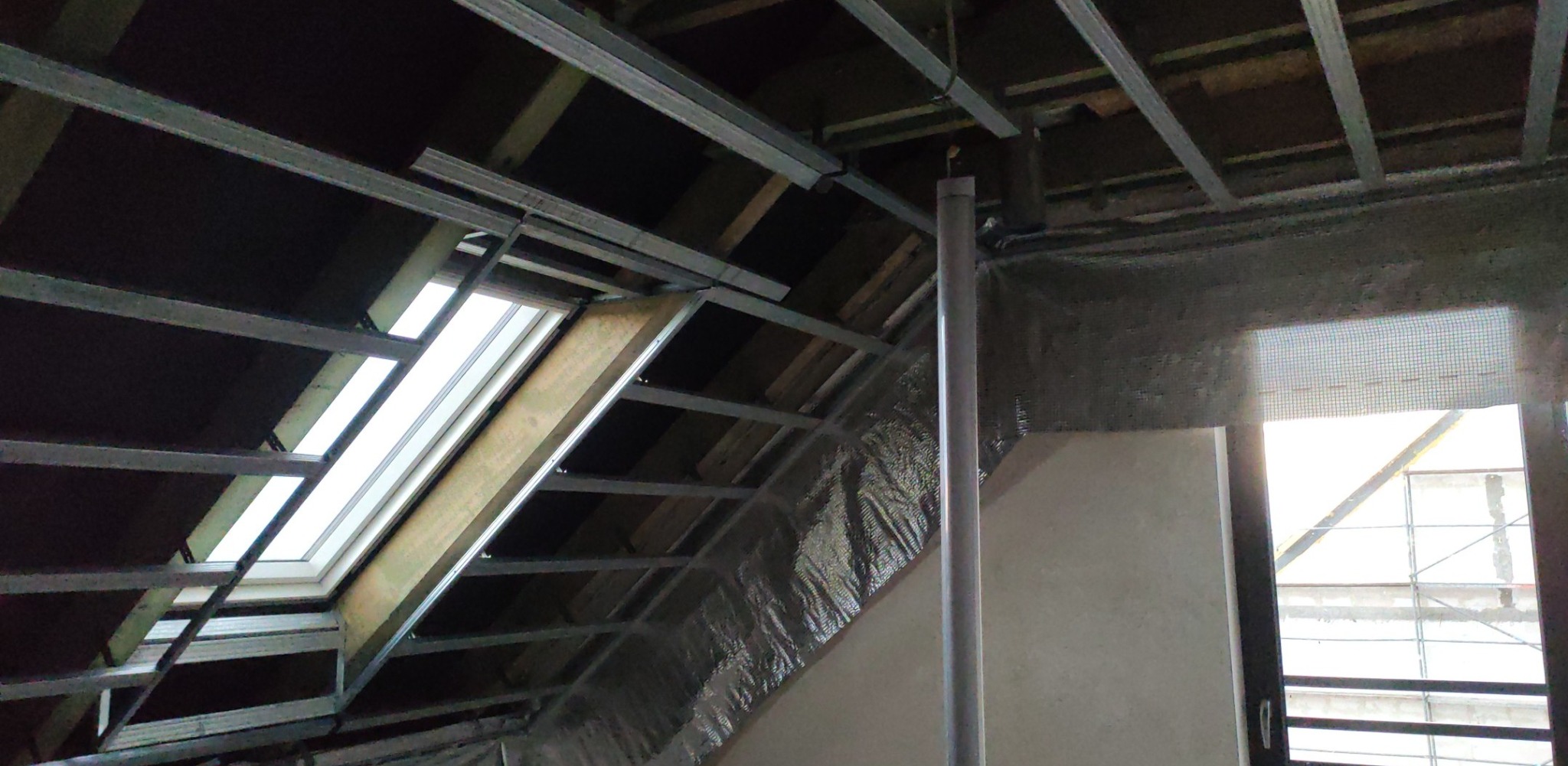 Montaż metalowej konstrukcji sufitu podwieszanego z oknem dachowym na poddaszu. Widoczne profile i folia izolacyjna na ścianach. Etap prac wykończeniowych.