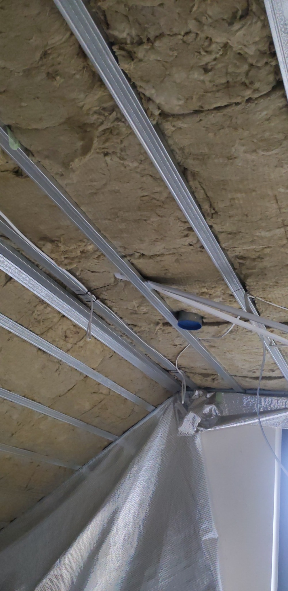 Montaż sufitu podwieszanego: widok na metalową konstrukcję z izolacją z wełny mineralnej, przewody elektryczne i fragment folii paroizolacyjnej przy ścianie.