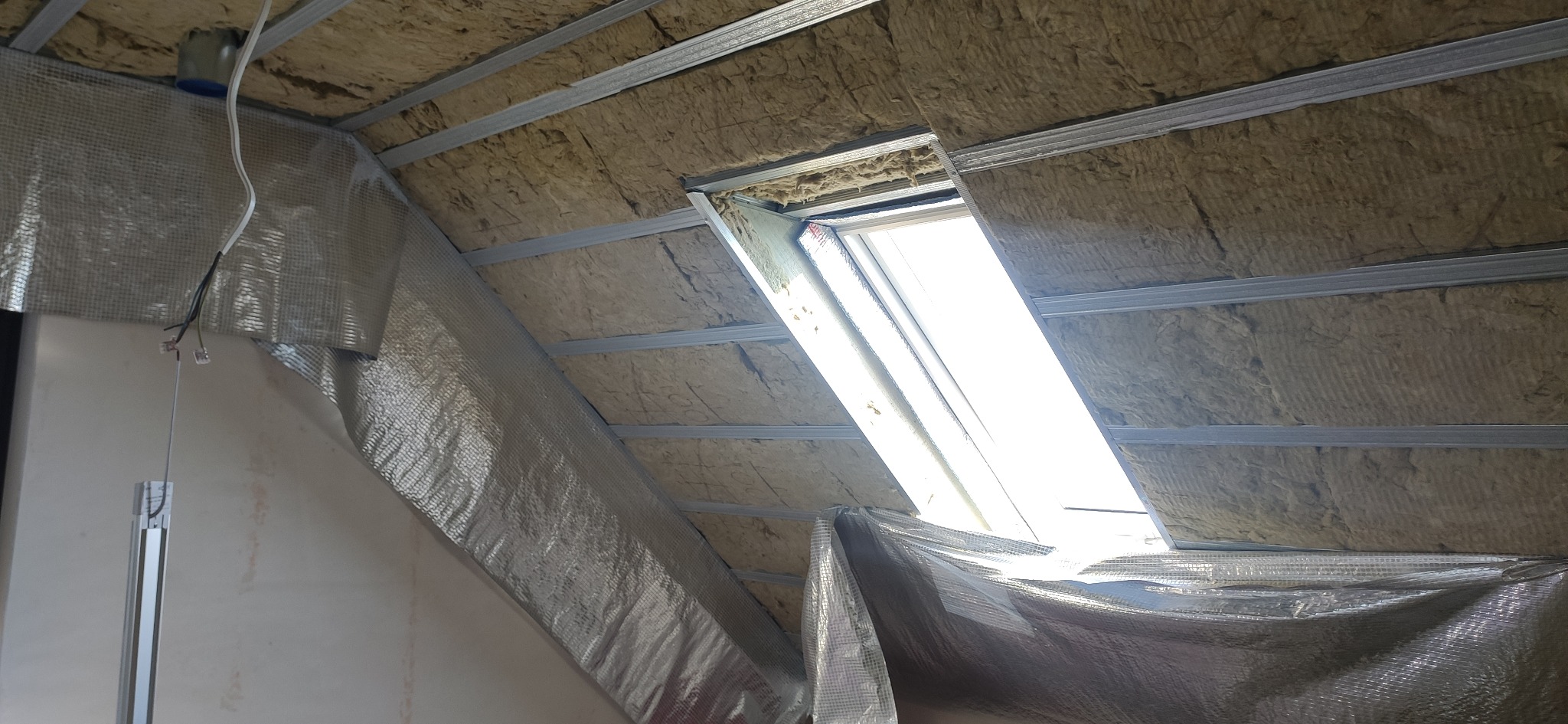 Izolacja poddasza wełną mineralną z folią aluminiową i metalowym stelażem, widoczne okno dachowe. Prace wykończeniowe na poddaszu.