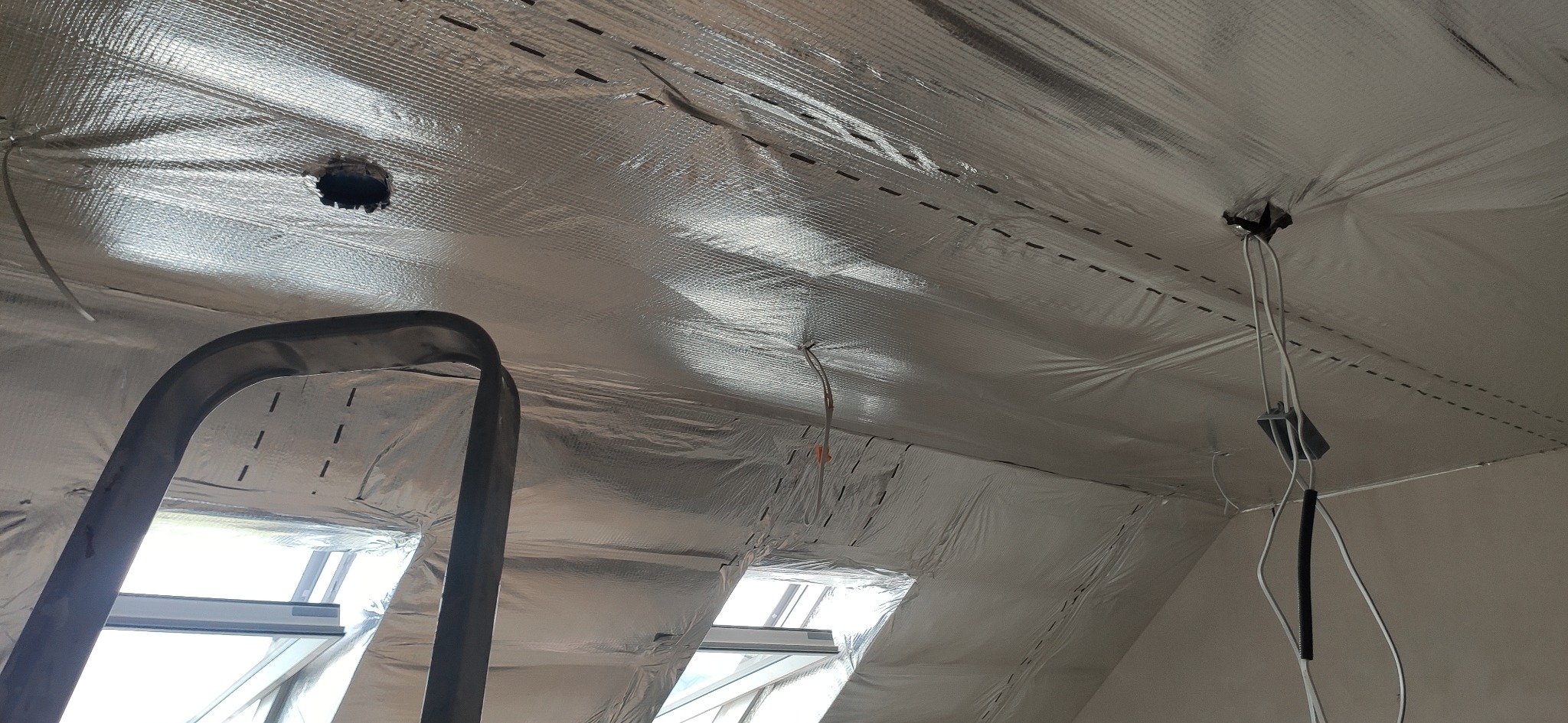 Izolacja poddasza z folią aluminiową, widoczne okna dachowe i drabina. Przygotowanie do wykończenia wnętrza, otwory na oświetlenie.