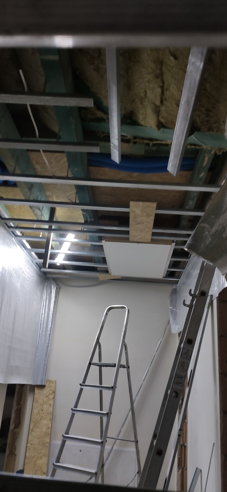 Montaż sufitu podwieszanego z płyt karton-gips, widoczne profile, izolacja i drabiny. Prace wykończeniowe w pomieszczeniu.