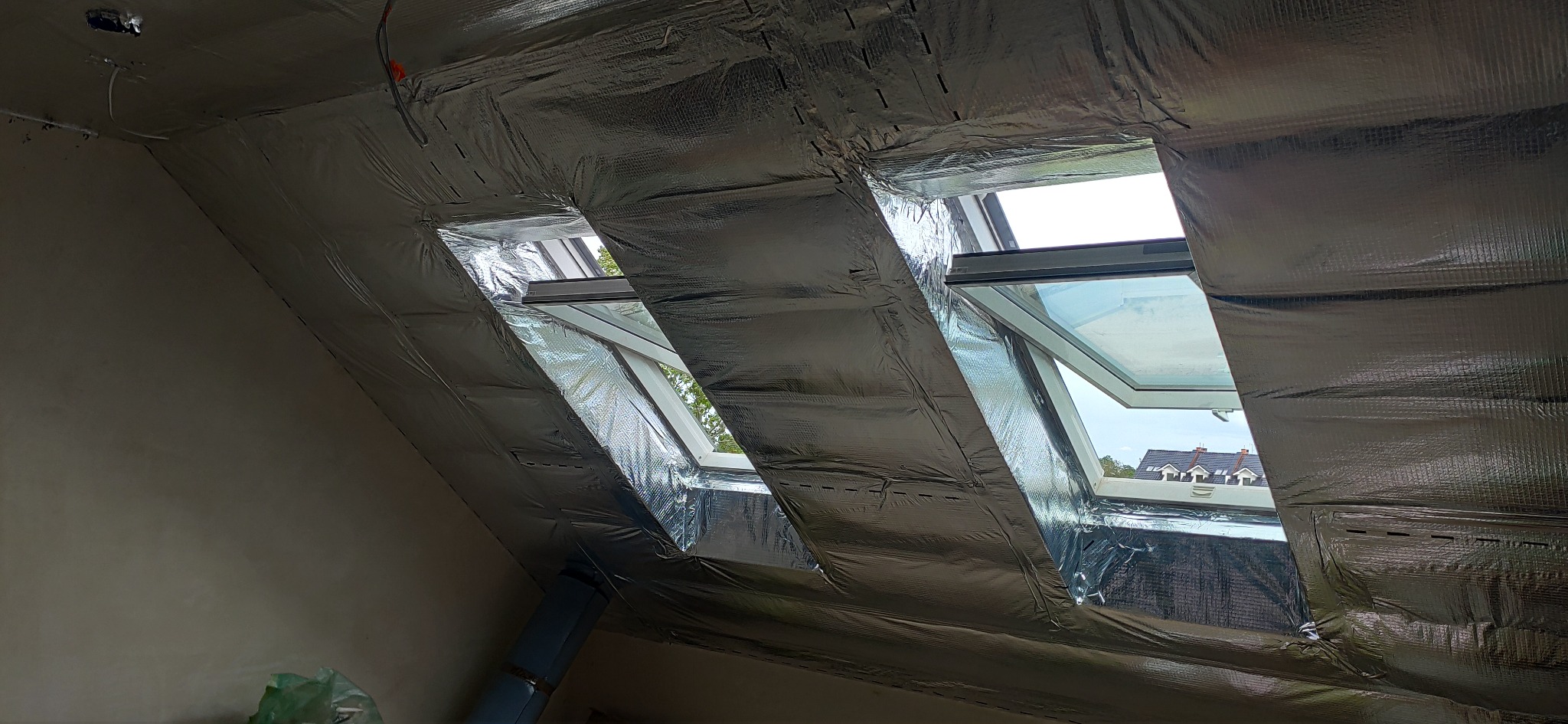 Izolacja poddasza folią aluminiową z trzema oknami dachowymi, widok na zewnątrz. Etap prac wykończeniowych, widoczne łączenia folii. Jasne światło dzienne.