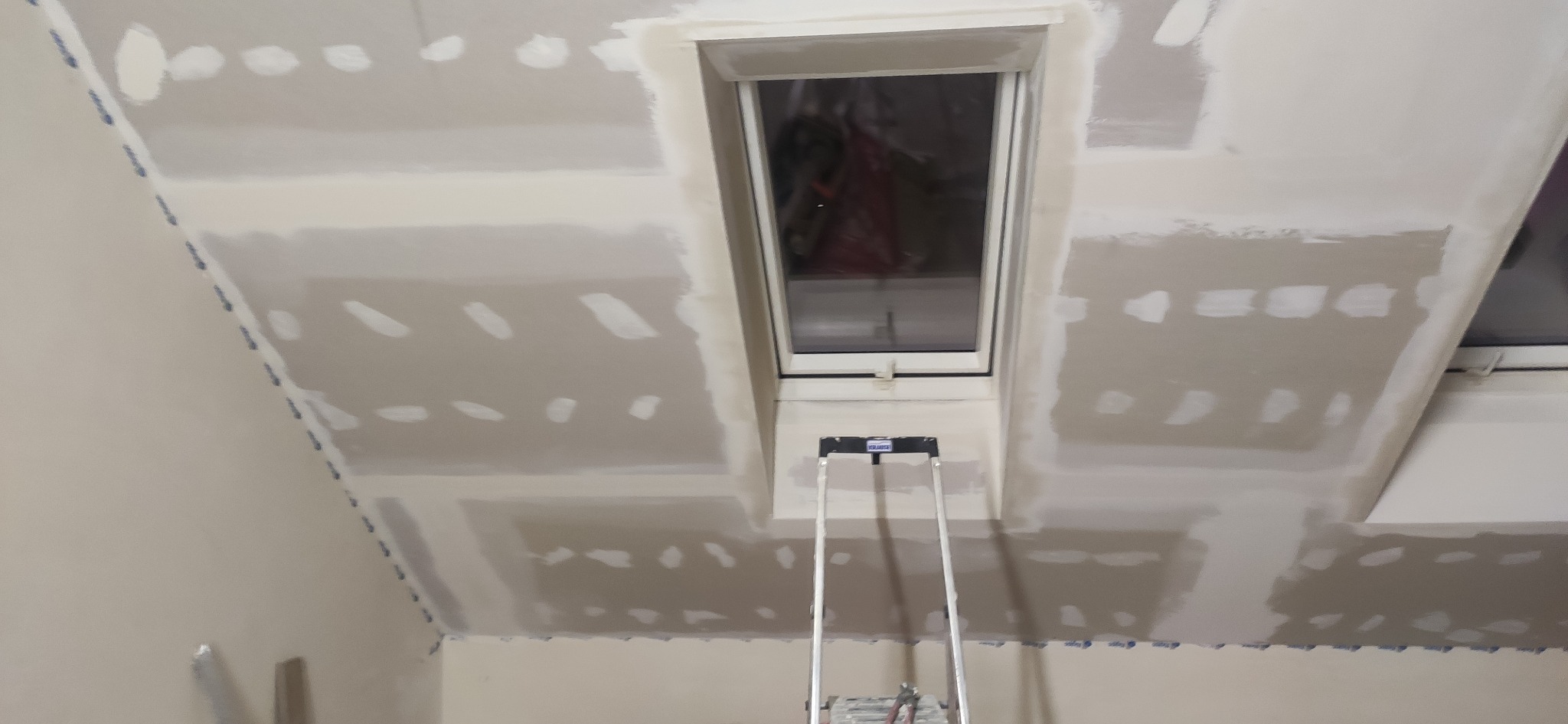 Poddasze z oknem dachowym w trakcie szpachlowania. Widoczne łączenia płyt gipsowych i drabina. Ściany i sufit przygotowane do dalszych prac wykończeniowych.