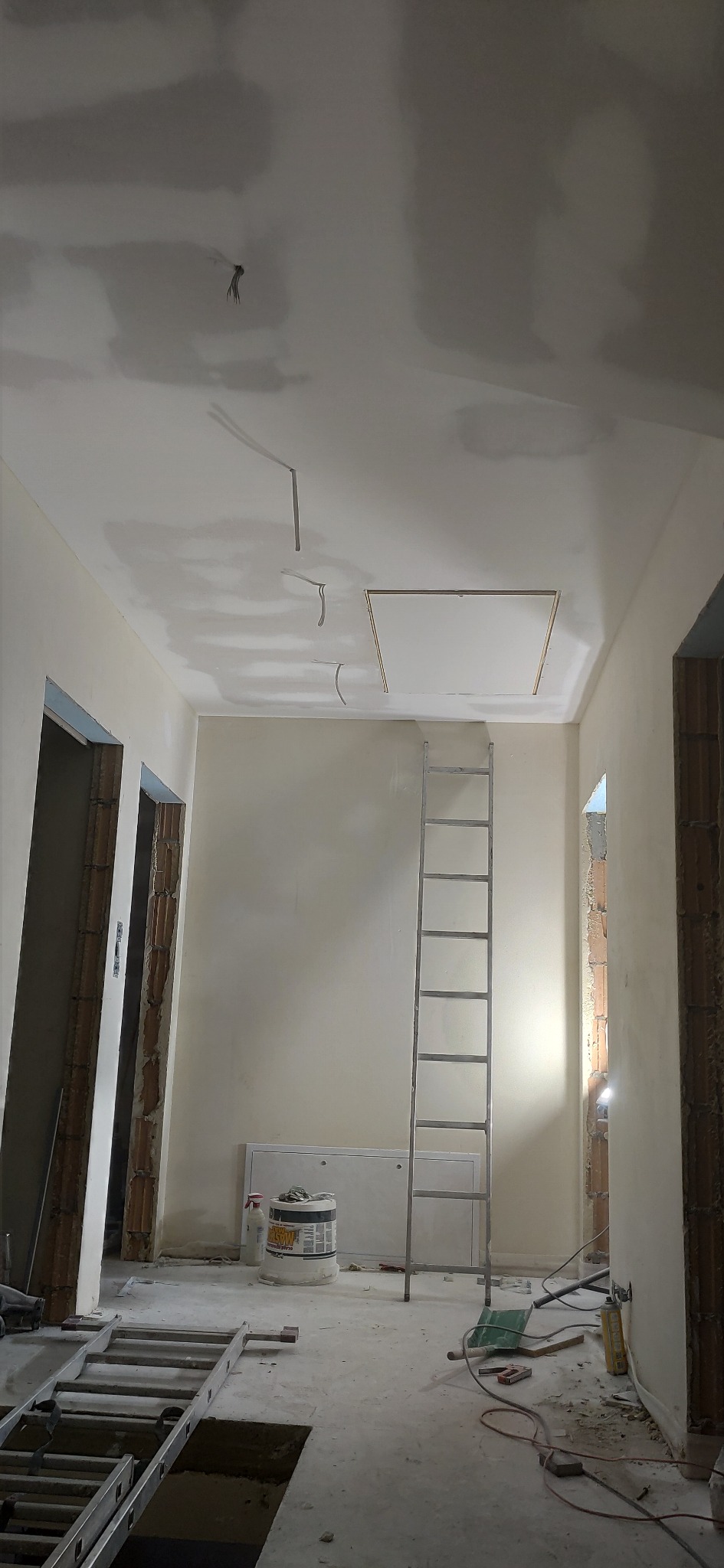 Wnętrze w trakcie remontu: ściany i sufit po szpachlowaniu, widoczna drabina, narzędzia i materiały budowlane. Otwory w ścianach przygotowane pod montaż ościeżnic.