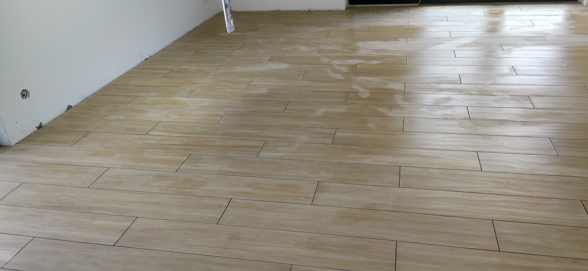 Ułożona podłoga z płytek imitujących drewno w jasnym odcieniu, widoczne fugi i fragment ściany z niedokończonym wykończeniem. Perspektywa z niskiego kąta.