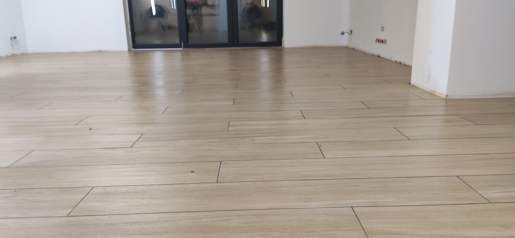 Ułożona podłoga z płytek imitujących drewno w jasnym odcieniu, widoczne fugi, ściany w kolorze białym, w tle przeszklone drzwi balkonowe.