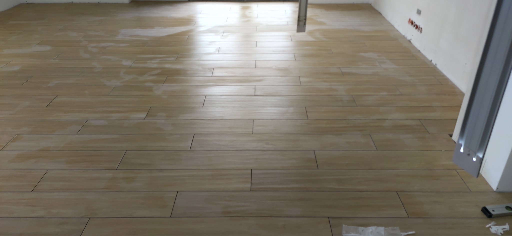Ułożona podłoga z płytek imitujących drewno w jasnym odcieniu, widoczne fugi i fragment ściany z instalacją elektryczną. W tle pionowa rura i fragment futryny.