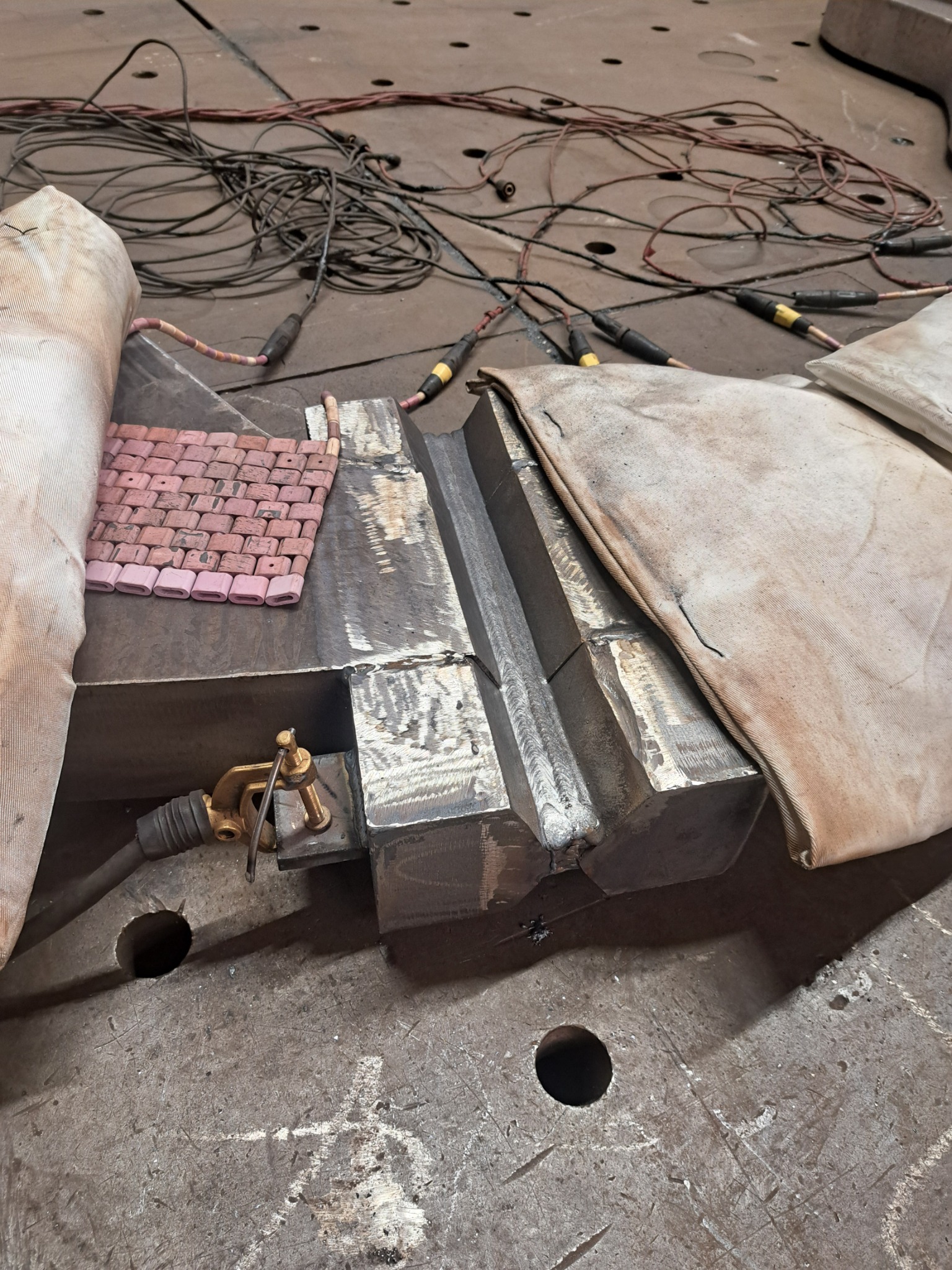 Metalowy element po obróbce, widoczne ślady spawania, osłony i narzędzia na metalowym stole z otworami. Kable i przewody w tle. Detal z bliska.