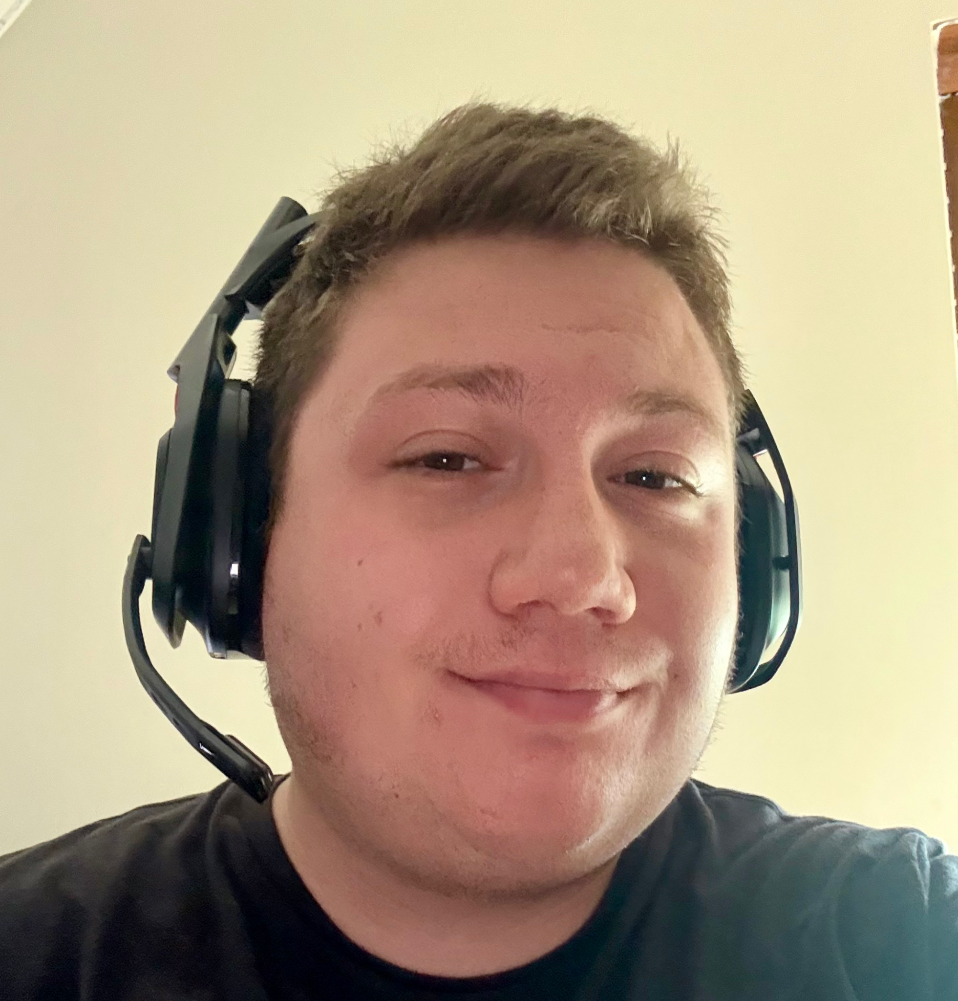 Mężczyzna w czarnej koszulce i słuchawkach na głowie, uśmiecha się patrząc w obiektyw, na jasnym tle. Widoczne detale słuchawek.