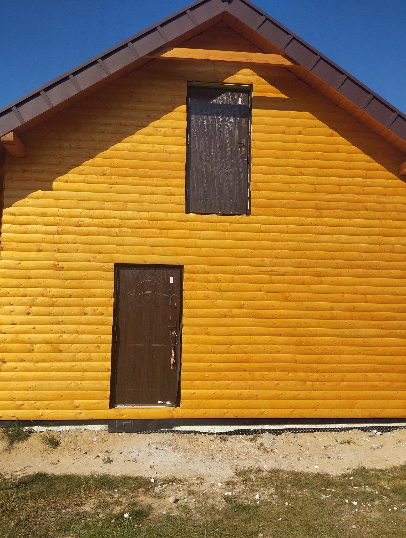 Drewniany dom z brązowymi drzwiami na parterze i poddaszu, widok z zewnątrz. Elewacja wykonana z drewnianych bali. Oświetlenie słoneczne podkreśla strukturę drewna.