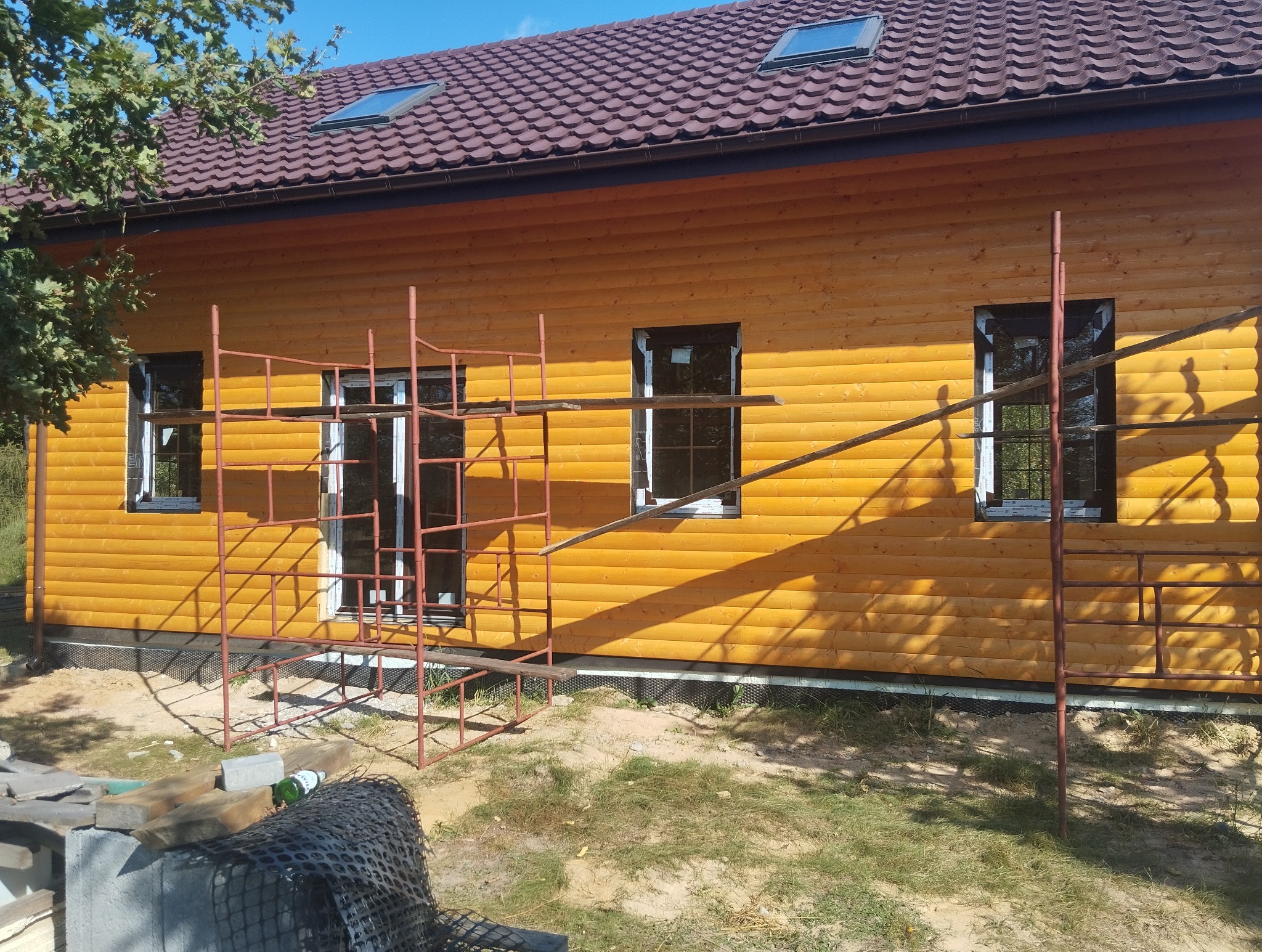 Dom z drewnianą elewacją w trakcie montażu, widoczne rusztowania i okna. Elewacja w ciepłym, złocistym odcieniu. Dach z brązowej blachodachówki.