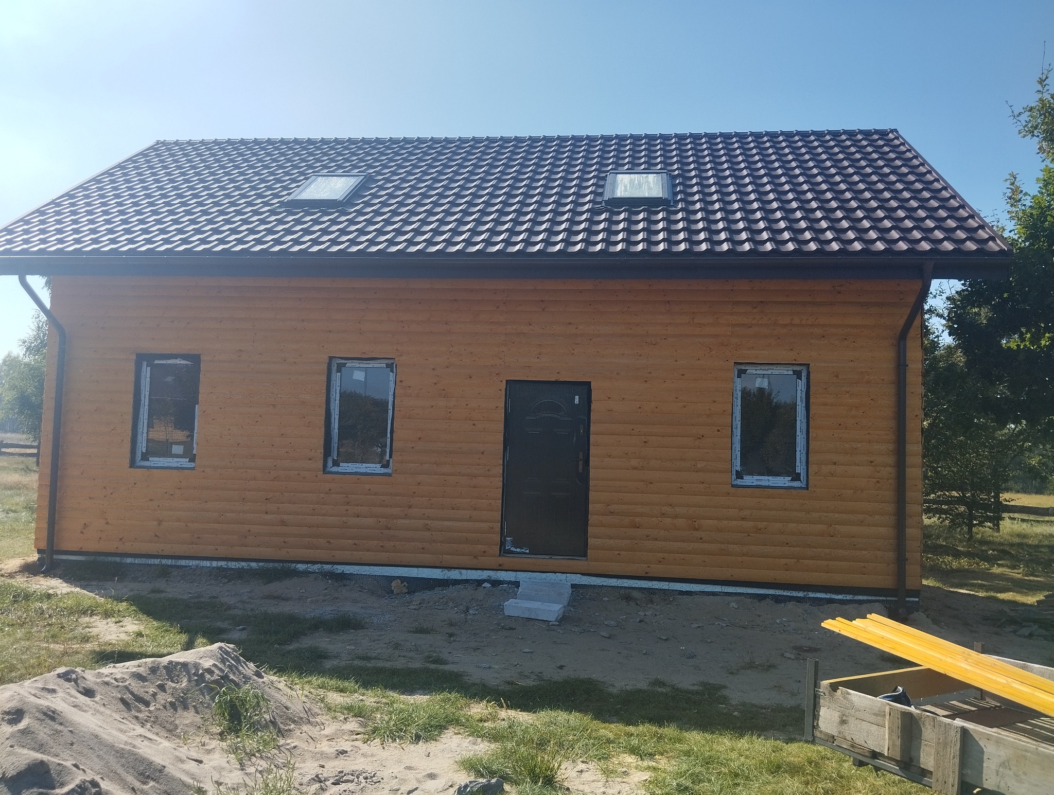 Drewniany dom z brązowym dachem i dwoma oknami dachowymi, świeżo po budowie, z widocznymi detalami wykończenia i elementami otoczenia w słoneczny dzień.