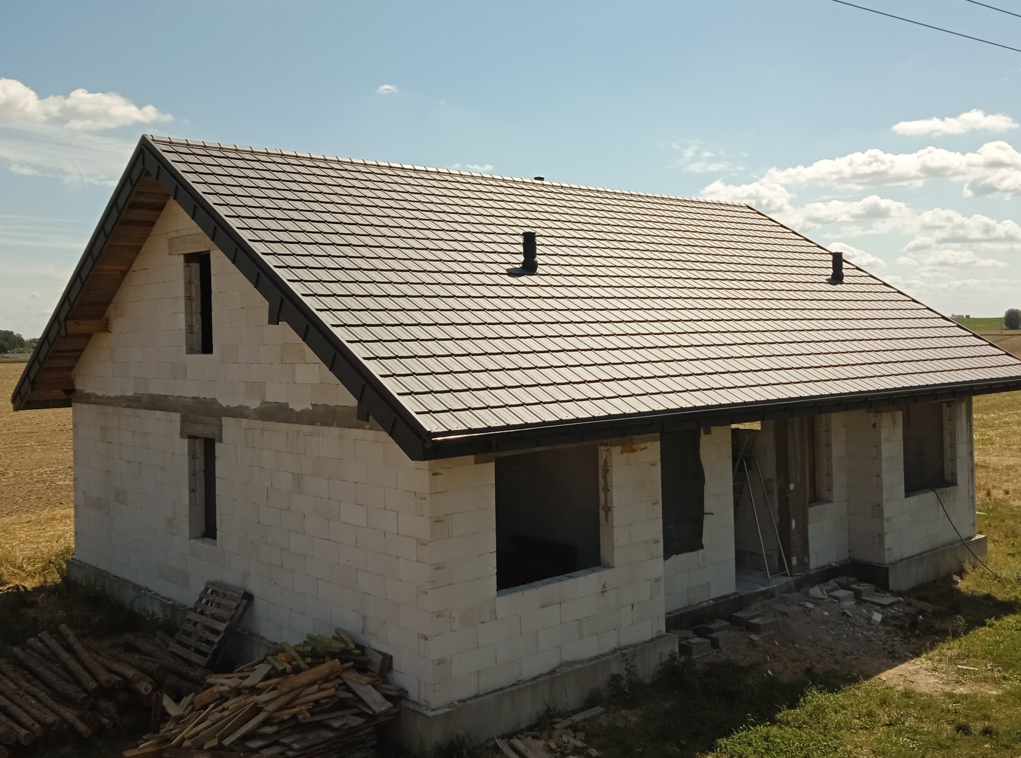 Surowy dom jednorodzinny z szarym, nowym dachem z dachówki ceramicznej. Widoczne kominy wentylacyjne. Ściany z białego betonu komórkowego. Na ziemi leżą deski.