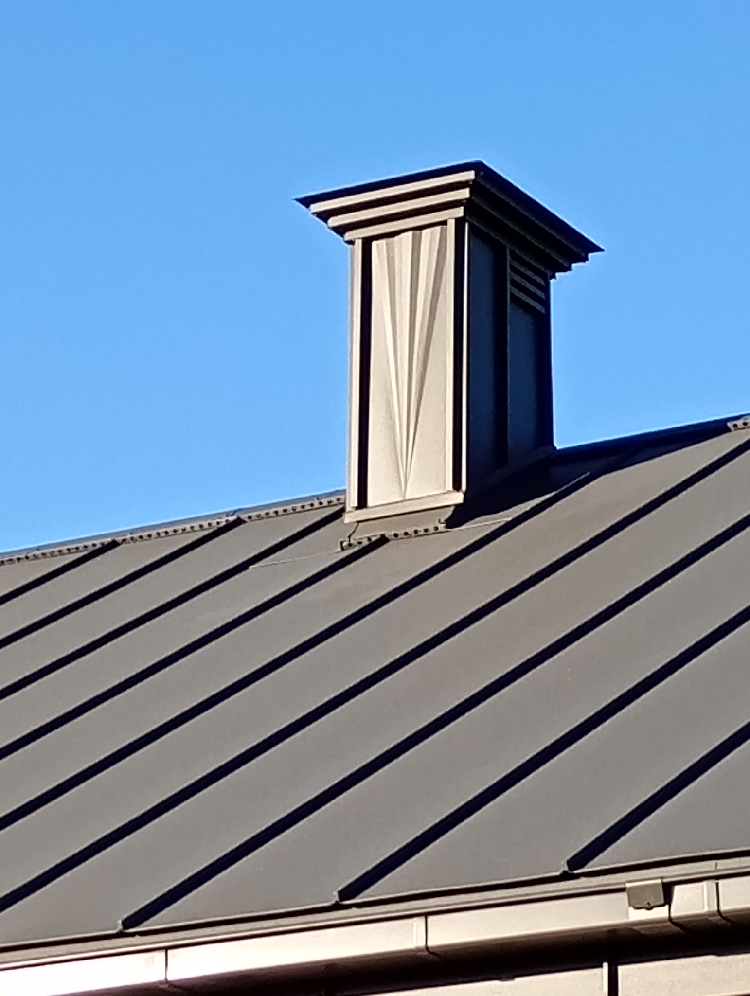 Szary dach z blachy trapezowej z ozdobnym kominem na tle błękitnego nieba. Widoczne mocowania blachy. Komin z dekoracyjnymi żłobieniami.