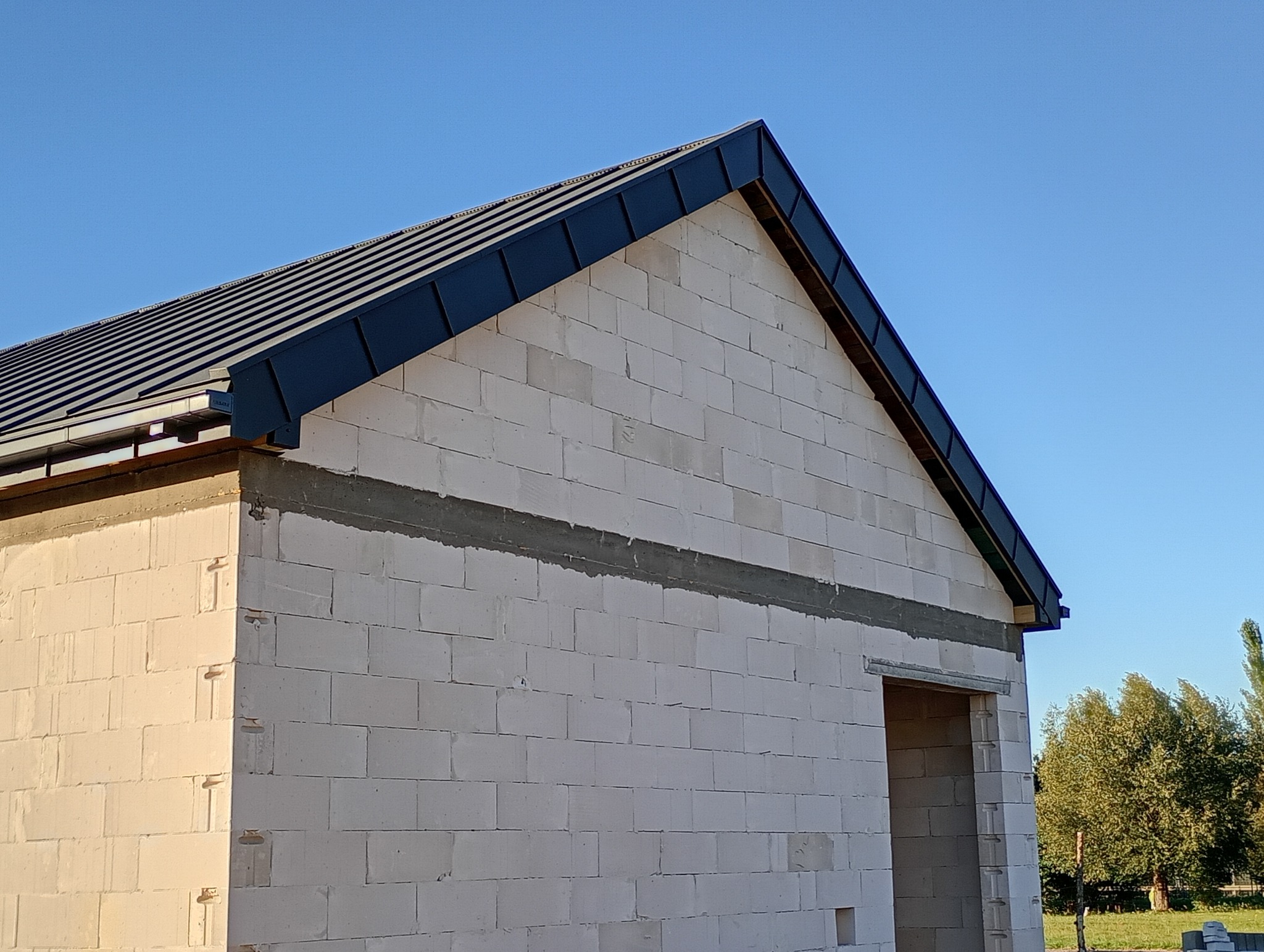 Surowy budynek z czarnym dachem z blachy na tle błękitnego nieba. Widoczna ściana z betonu komórkowego i otwór drzwiowy. Budowa w toku, etap krycia dachu.