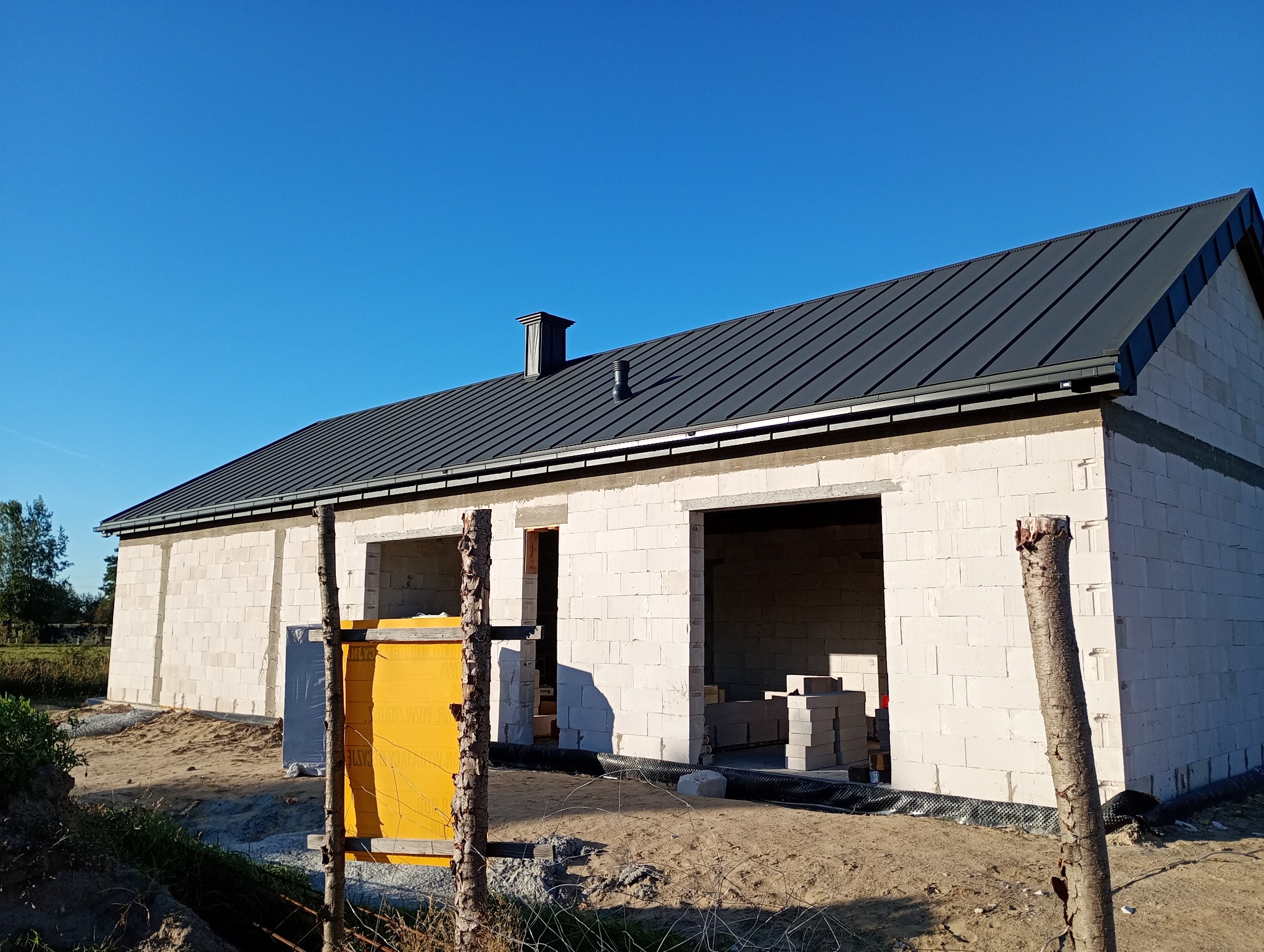 Dom w trakcie budowy z szarą blachą na dachu i białymi ścianami z betonu komórkowego. Widoczne materiały izolacyjne i elementy konstrukcyjne.