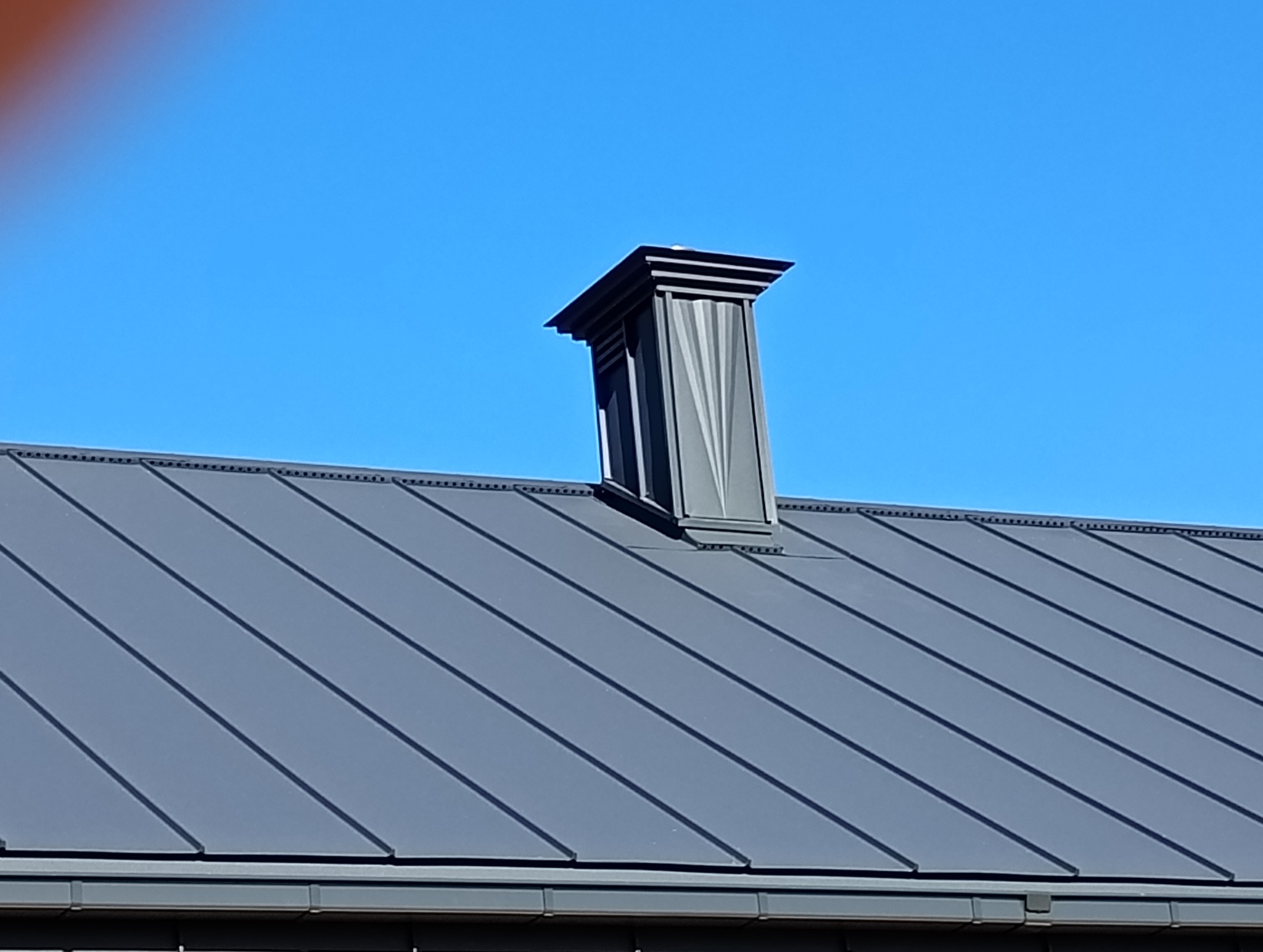 Szary dach z blachy trapezowej z ozdobnym kominem na tle błękitnego nieba. Widoczne łączenia blachy i detale komina. Ujęcie z dołu, podkreślające geometrię dachu.