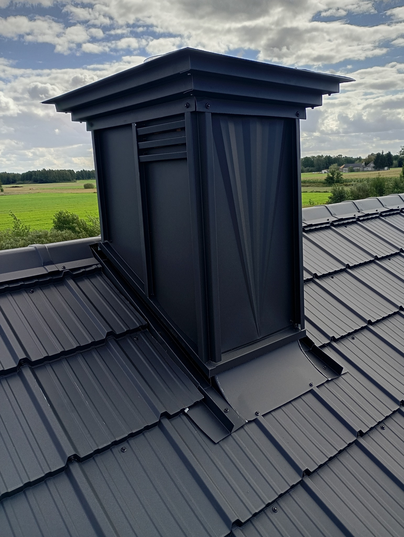Czarny komin wentylacyjny na dachu z blachy trapezowej, widok z góry, na tle błękitnego nieba z chmurami i zielonego krajobrazu. Detal wykończenia dachu.