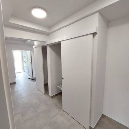 Kamil Kaczmarczyk PrimeHome - Jasne wnętrze z przesuwnymi drzwiami w minimalistycznym stylu. Szara podłoga z płytek imitujących kamień. Widok na korytarz i drzwi wejściowe.