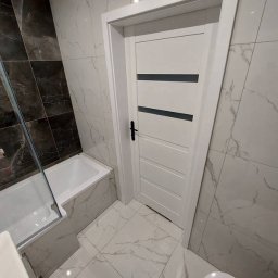 Kamil Kaczmarczyk PrimeHome - Nowoczesna łazienka z biało-szarymi marmurkowymi płytkami, białą wanną i drzwiami z czarnymi akcentami. Minimalistyczny design, oświetlenie punktowe.