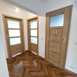 Kamil Kaczmarczyk PrimeHome - Korytarz z trzema drewnianymi drzwiami z przeszkleniami, parkiet ułożony w jodełkę. Jedne drzwi oznaczone symbolem toalety damsko-męskiej. Nowoczesny design.