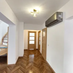 Korytarz z parkietem w jodełkę, drewnianymi drzwiami i klimatyzatorem na ścianie. Widoczne fragmenty schodów. Ciepłe oświetlenie.