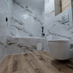 Kamil Kaczmarczyk PrimeHome - Wykończona łazienka z marmurowymi płytkami na ścianach i drewnopodobną podłogą. Wisząca toaleta i bateria wannowa w kolorze czarnym.