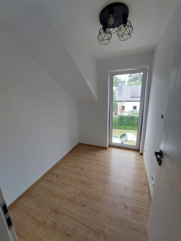 Puste, odświeżone wnętrze pokoju z oknem, panelami podłogowymi i nowoczesną lampą sufitową. Ściany pomalowane na biało, widoczny skos dachu.