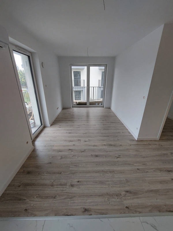 Jasne wnętrze z panelami podłogowymi imitującymi drewno. Widoczne okno balkonowe i drzwi balkonowe. Ściany pomalowane na biało, minimalistyczny styl wykończenia.