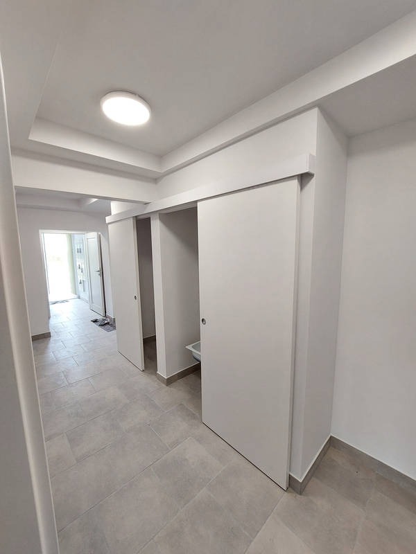 Jasne wnętrze z przesuwnymi drzwiami w minimalistycznym stylu. Szara podłoga z płytek imitujących kamień. Widok na korytarz i drzwi wejściowe.