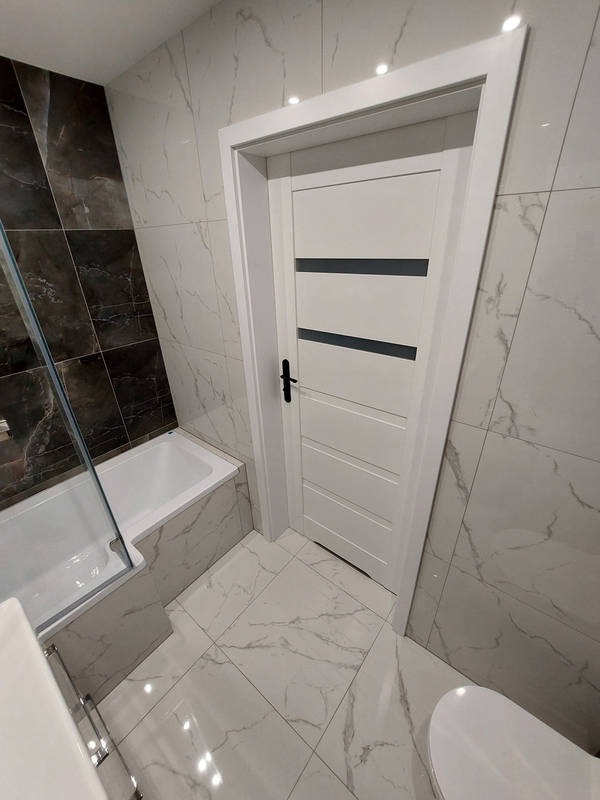 Nowoczesna łazienka z biało-szarymi marmurkowymi płytkami, białą wanną i drzwiami z czarnymi akcentami. Minimalistyczny design, oświetlenie punktowe.