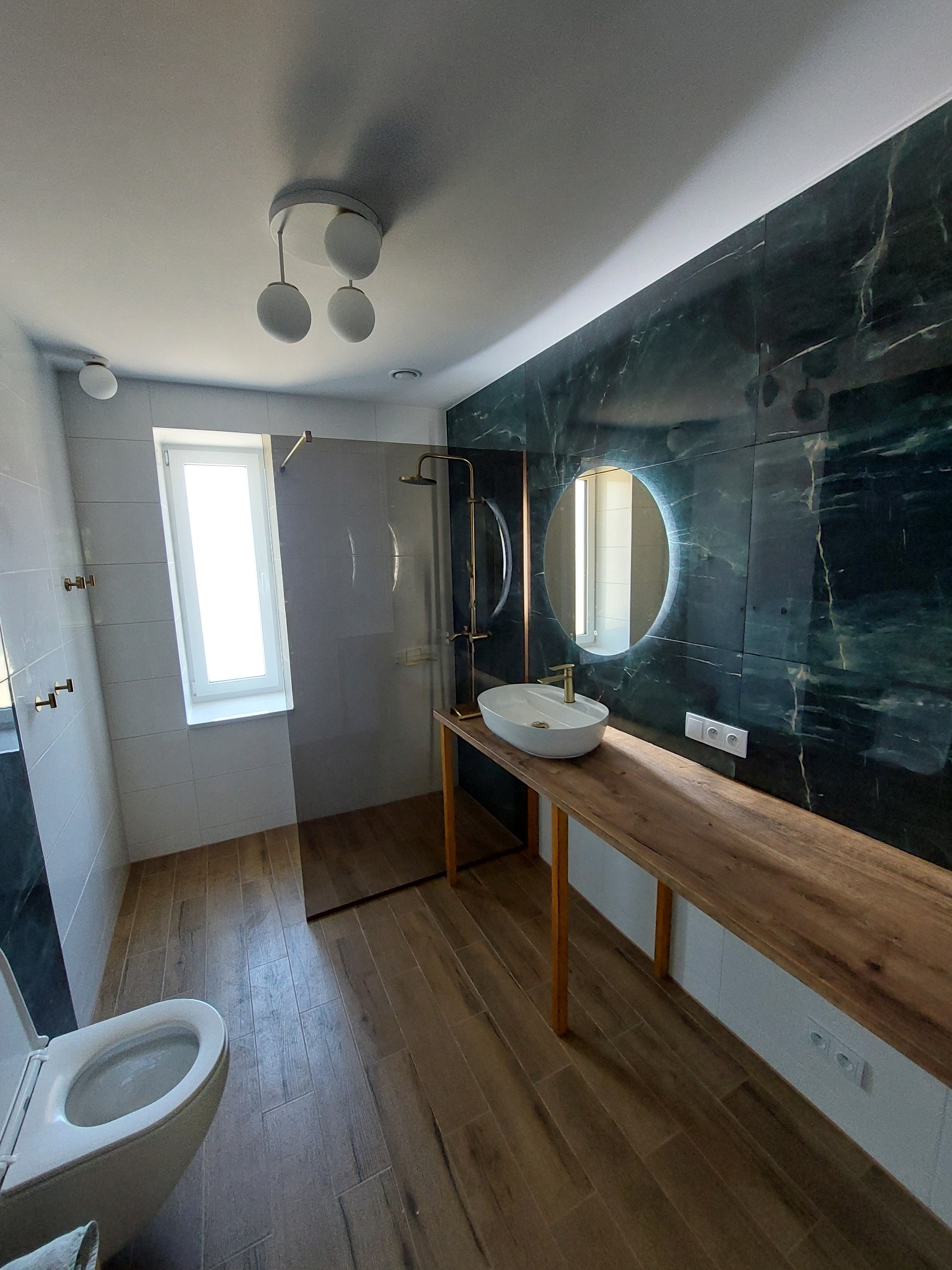 Elegancka łazienka z kabiną prysznicową, umywalką na drewnianym blacie i marmurową ścianą z okrągłym lustrem z podświetleniem LED. Jasne płytki i drewniana podłoga.