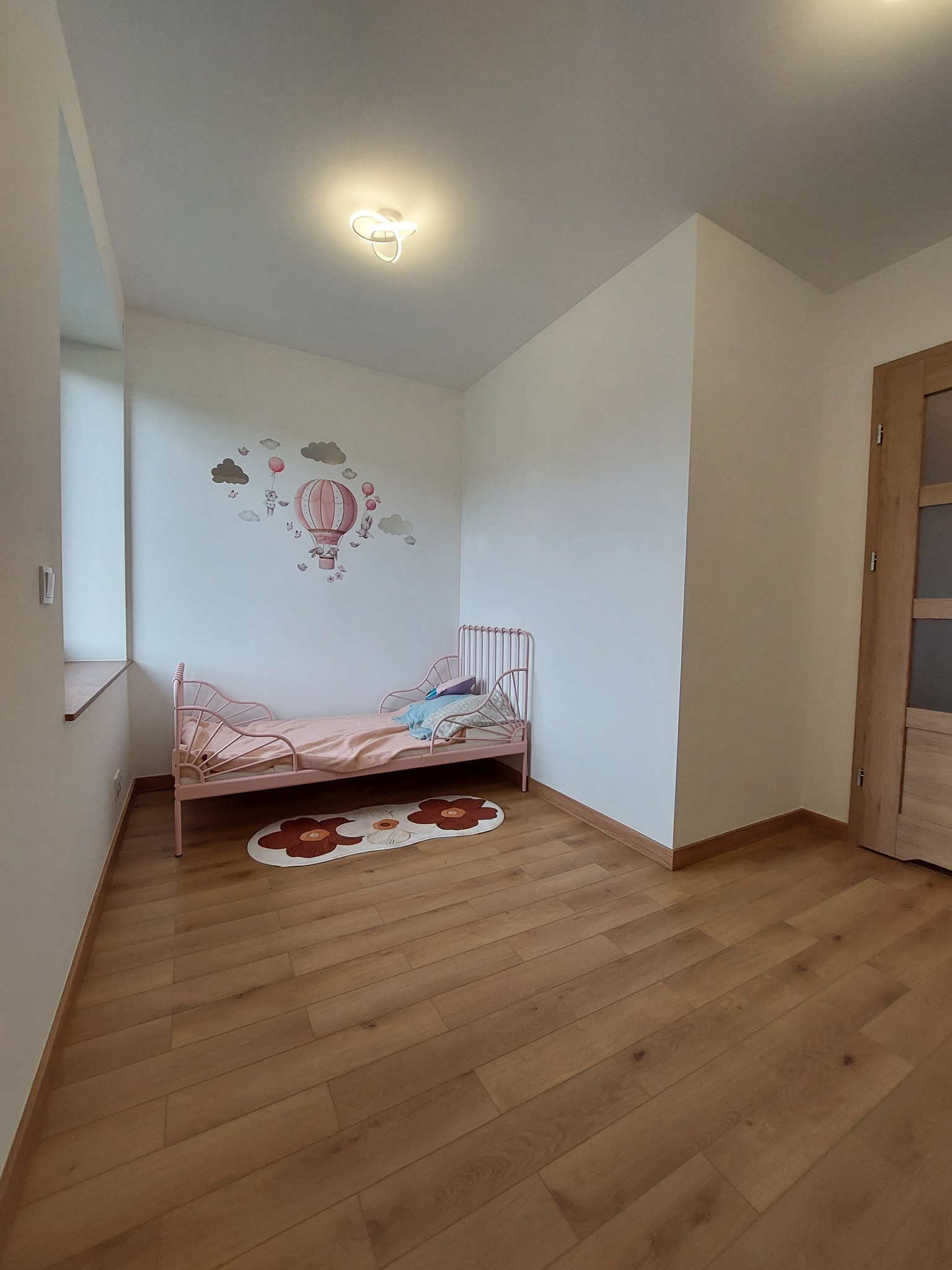 Pokój dziecięcy z drewnianą podłogą i różowym łóżeczkiem. Na ścianie naklejka z motywem balonu. Widoczne listwy przypodłogowe i drzwi z przeszkleniem.
