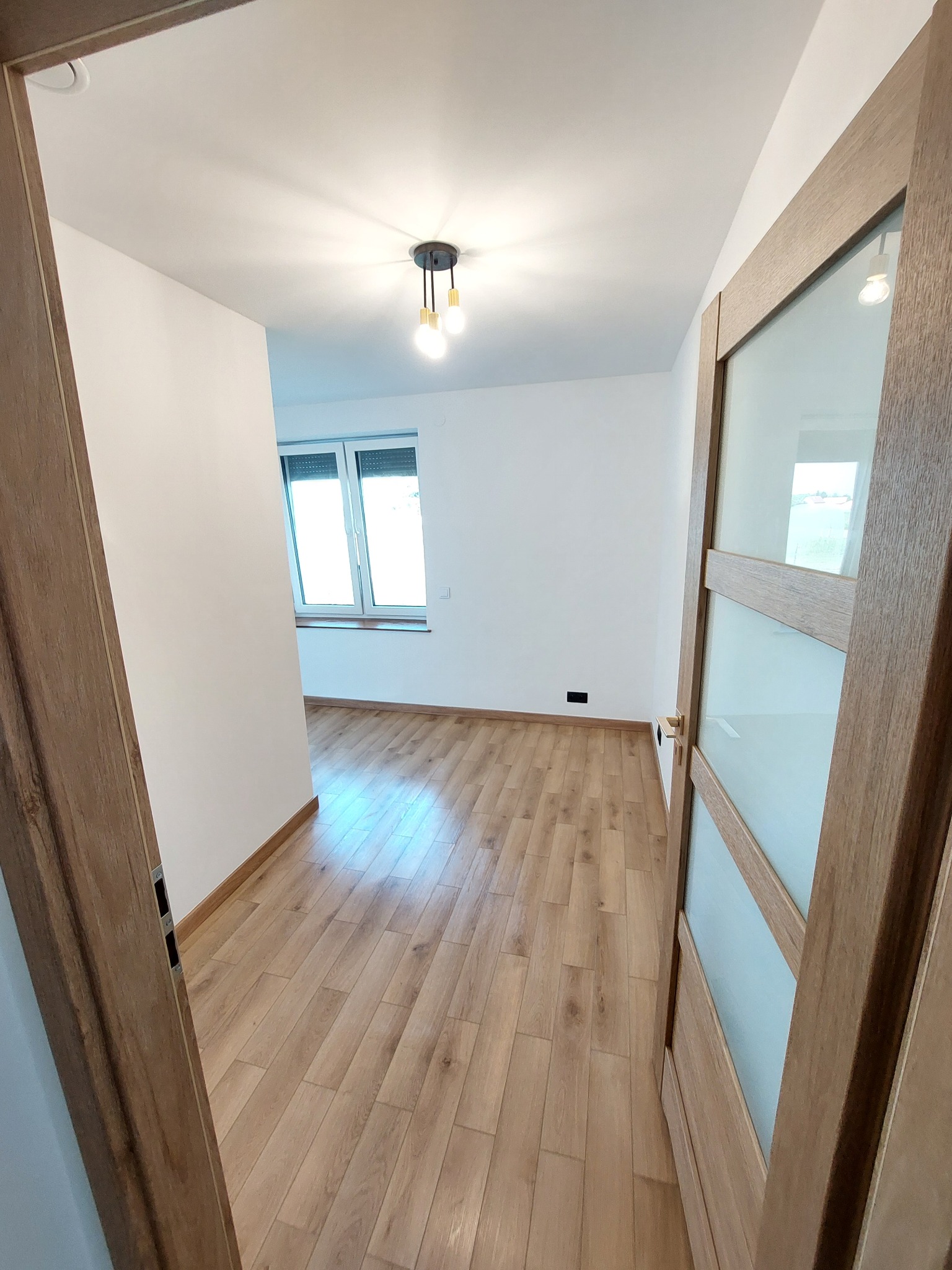 Jasne, odnowione wnętrze z drewnianą podłogą i drzwiami z przeszkleniami. Widok z perspektywy wejścia, minimalistyczne oświetlenie sufitowe.