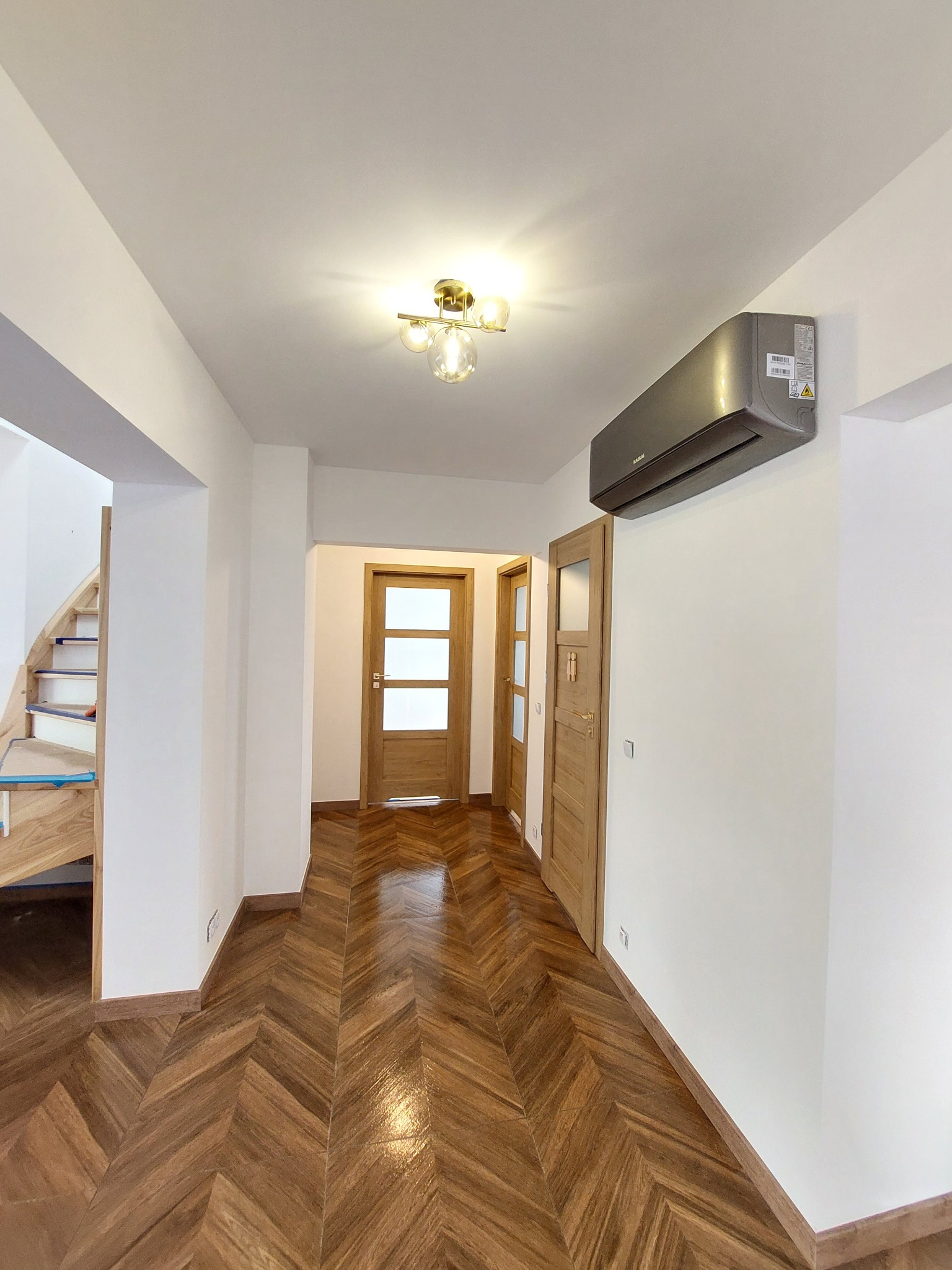 Korytarz z parkietem w jodełkę, drewnianymi drzwiami i klimatyzatorem na ścianie. Widoczne fragmenty schodów. Ciepłe oświetlenie.