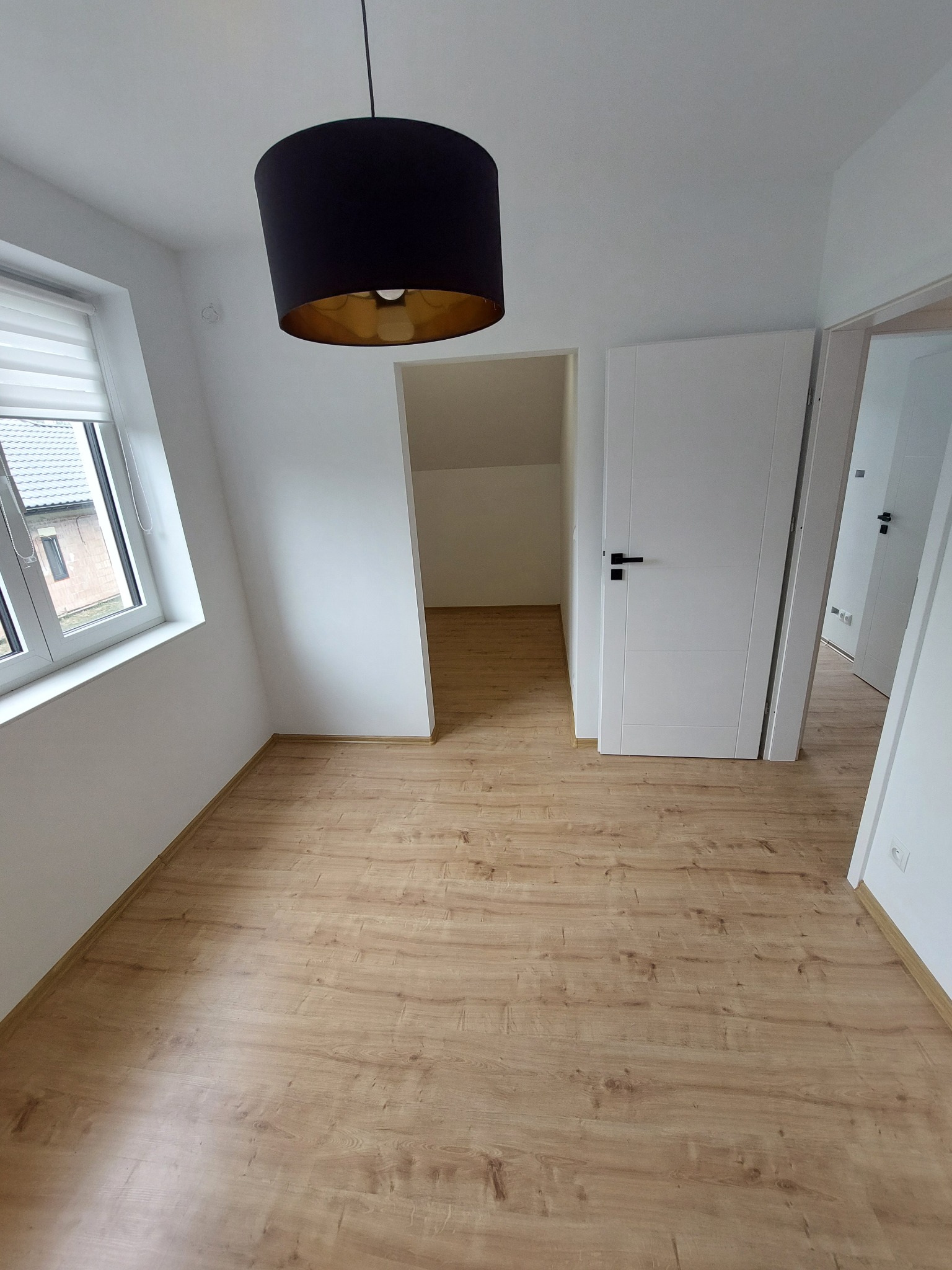 Jasne wnętrze po remoncie: panele podłogowe, białe ściany i drzwi z czarnymi klamkami. W oknie widoczne rolety. Czarna lampa z elementami złota.