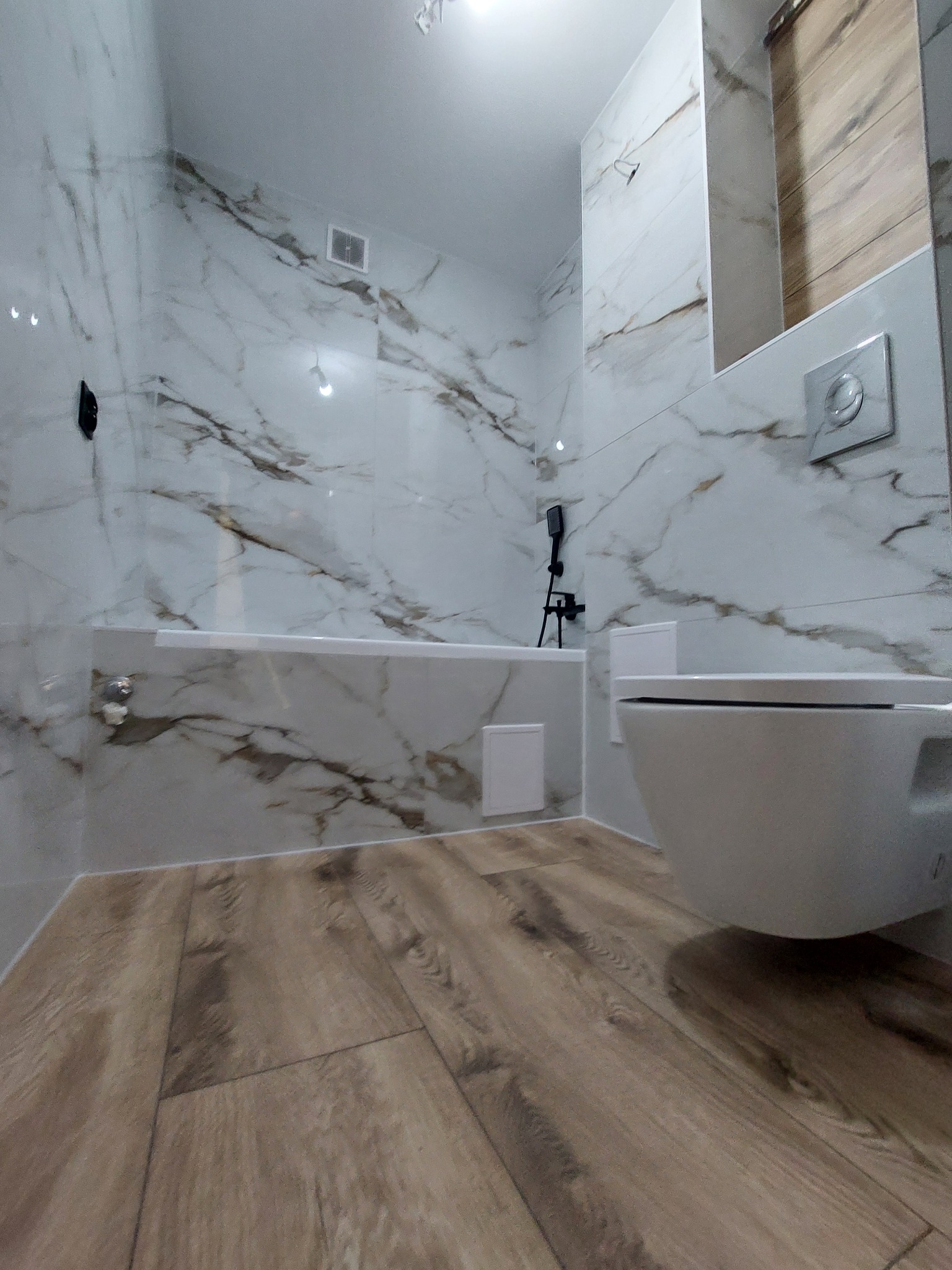 Wykończona łazienka z marmurowymi płytkami na ścianach i drewnopodobną podłogą. Wisząca toaleta i bateria wannowa w kolorze czarnym.