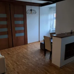 Salon z parkietem w jodełkę, drewnianymi drzwiami przesuwnymi i designerską lampą. Widoczne elementy dekoracyjne i meble w stonowanej kolorystyce.