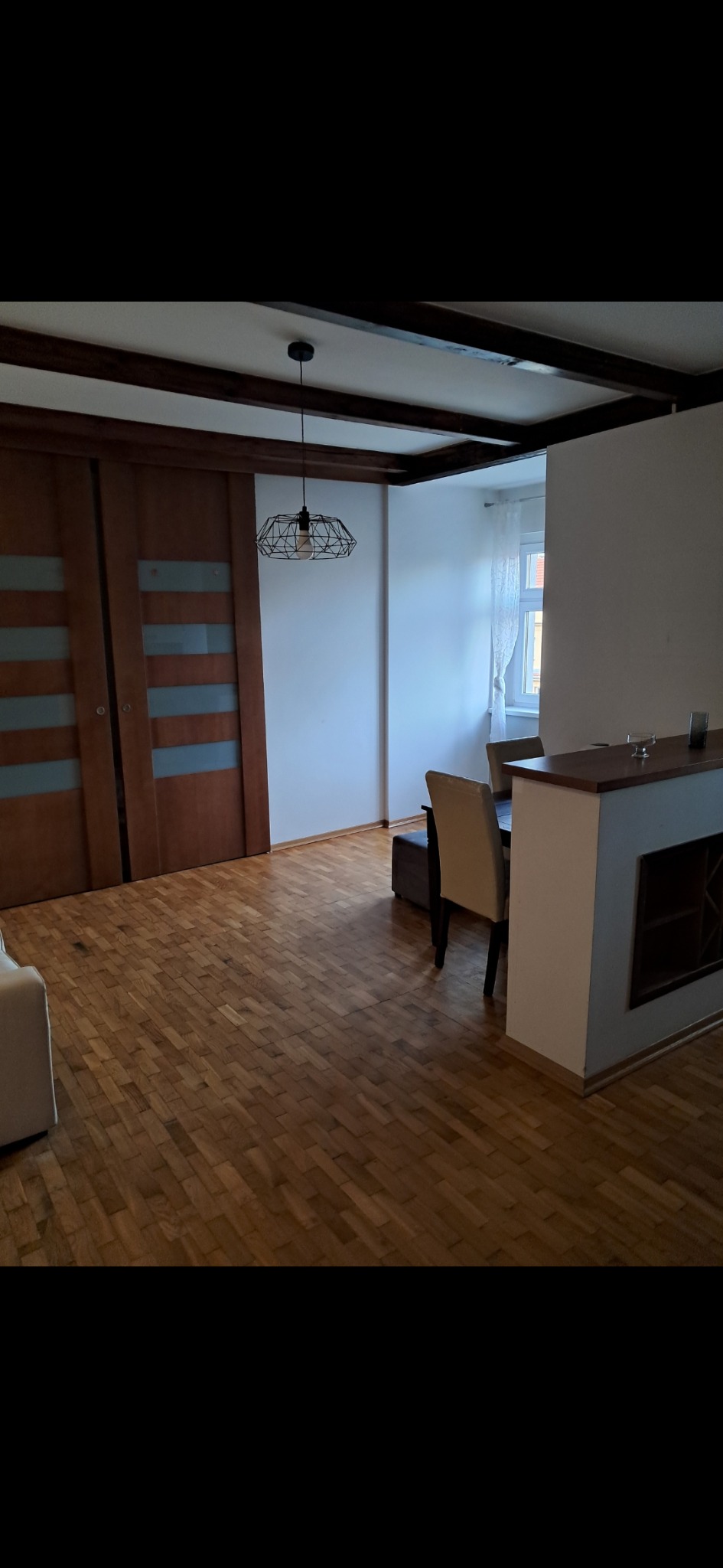 Salon z parkietem w jodełkę, drewnianymi drzwiami przesuwnymi i designerską lampą. Widoczne elementy dekoracyjne i meble w stonowanej kolorystyce.