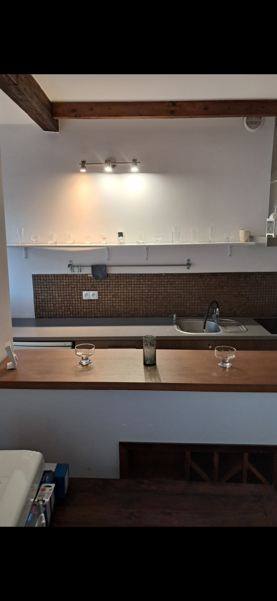 Kuchnia z drewnianym blatem i rzędem szklanek na półce. Mozaika na ścianie, lampa sufitowa. Umywalka i bateria kuchenna. Minimalistyczny wystrój wnętrza.