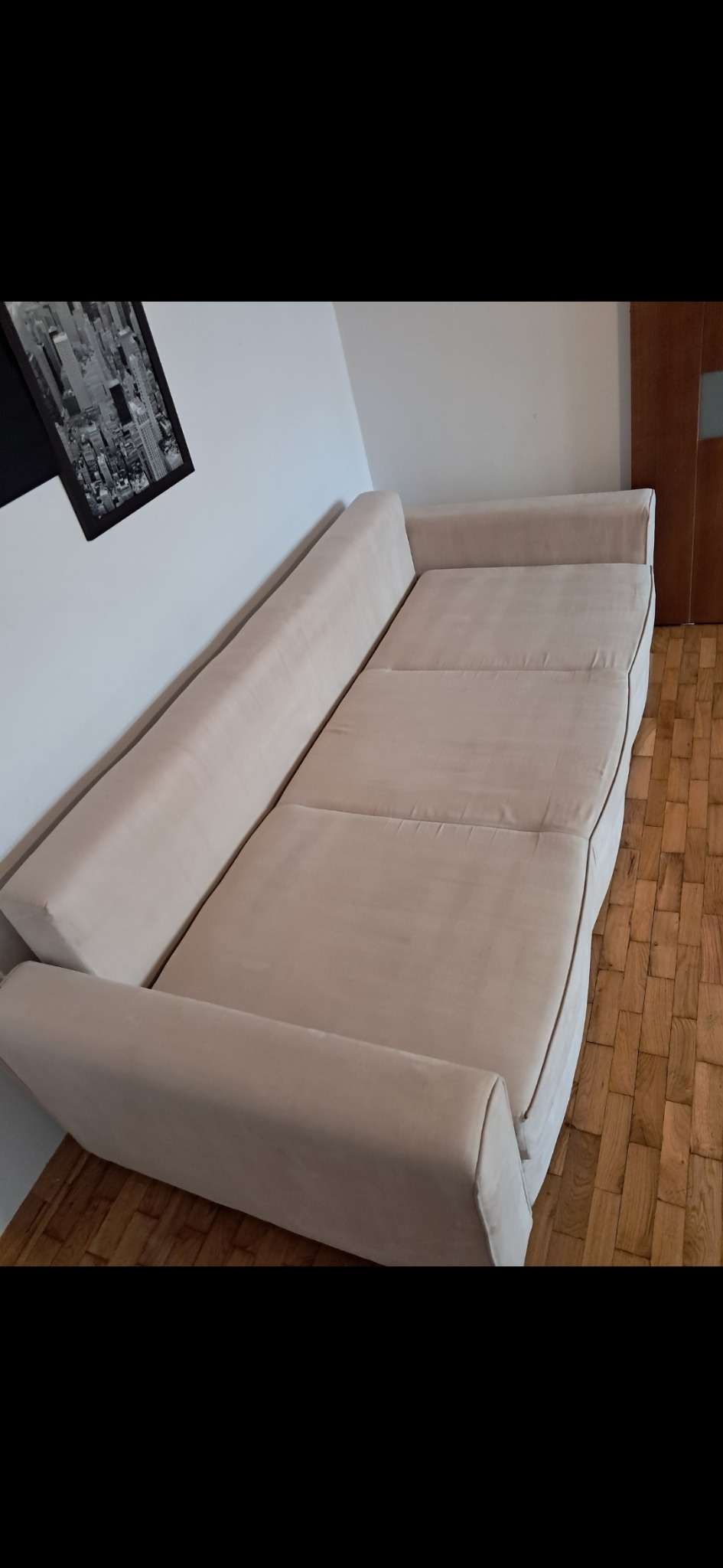 Rozłożona, beżowa kanapa z poduszkami na drewnianej podłodze, widziana z boku. Na ścianie wisi czarno-biały obraz w ramie.