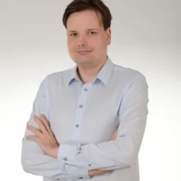 Portret mężczyzny w jasnej koszuli z podwiniętymi rękawami, stojącego na jasnym tle z założonymi rękami. Uśmiech na twarzy, naturalne światło.