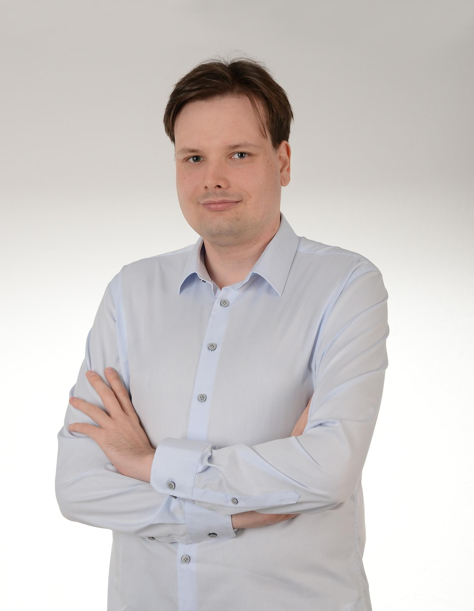 Portret mężczyzny w jasnej koszuli z podwiniętymi rękawami, stojącego na jasnym tle z założonymi rękami. Uśmiech na twarzy, naturalne światło.